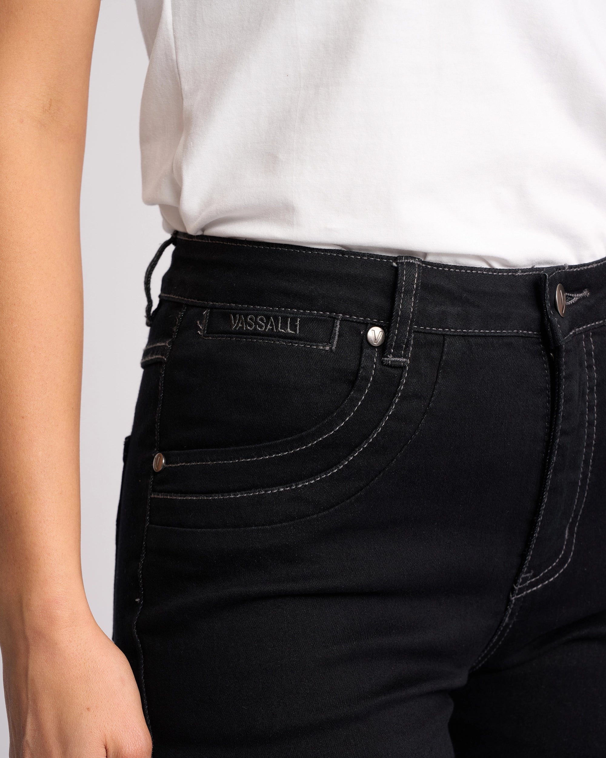 A person wears a white shirt and Vassalli Studio Jean - Black Denim featuring visible white stitching and a VASSALLI label above the front pocket. Their left hand rests relaxed by their side.