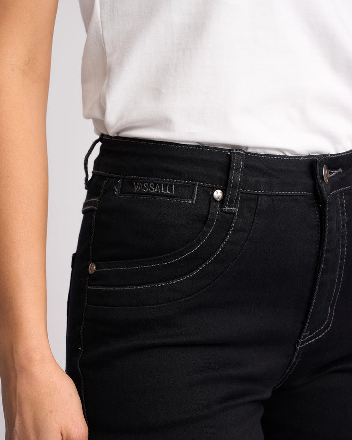 A person wears a white shirt and Vassalli Studio Jean - Black Denim featuring visible white stitching and a VASSALLI label above the front pocket. Their left hand rests relaxed by their side.