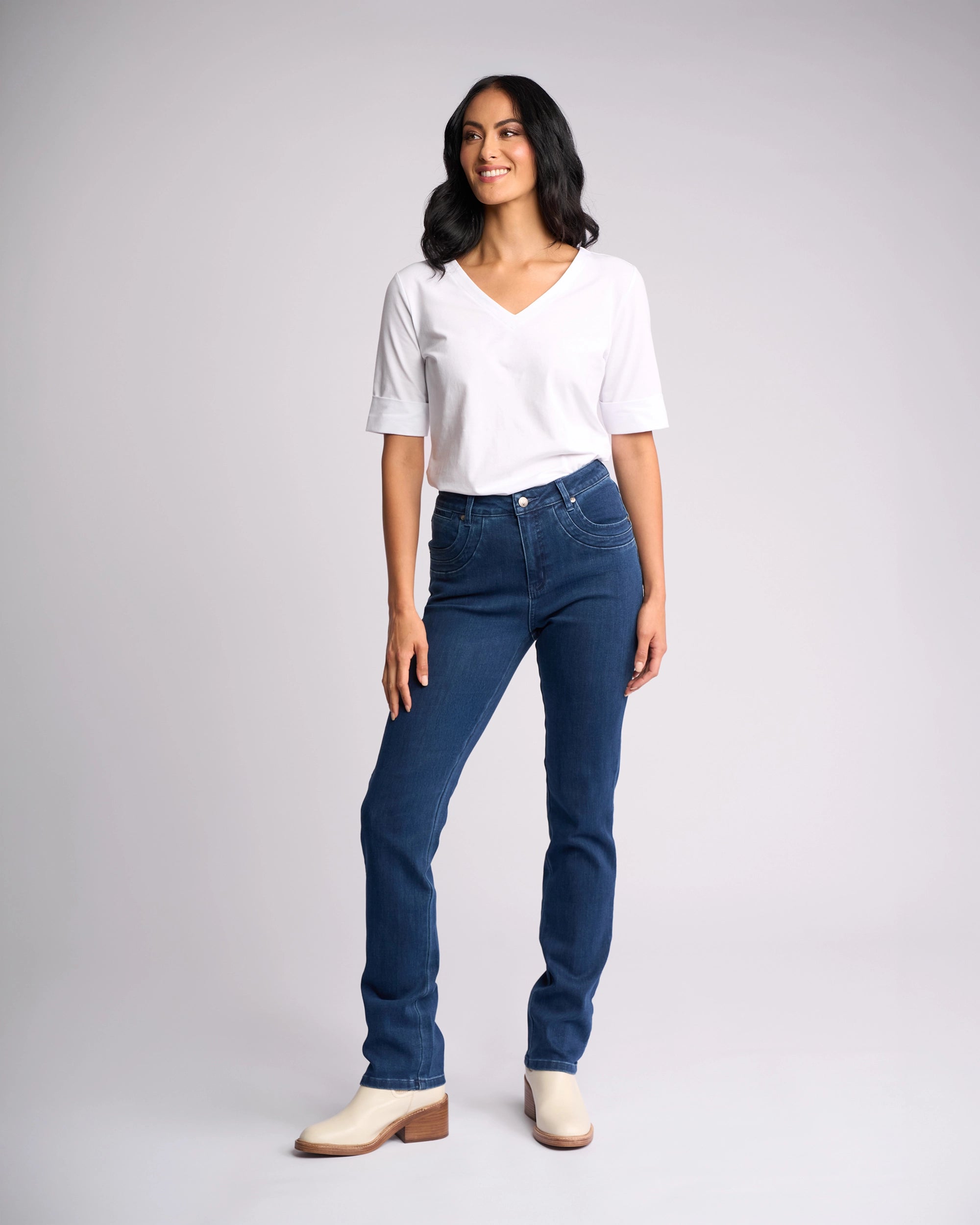 A woman with dark, shoulder-length hair smiles while wearing Vassalli’s Studio Jean in blue denim and a white V-neck tee, paired with cream ankle boots, standing against a plain light gray background.