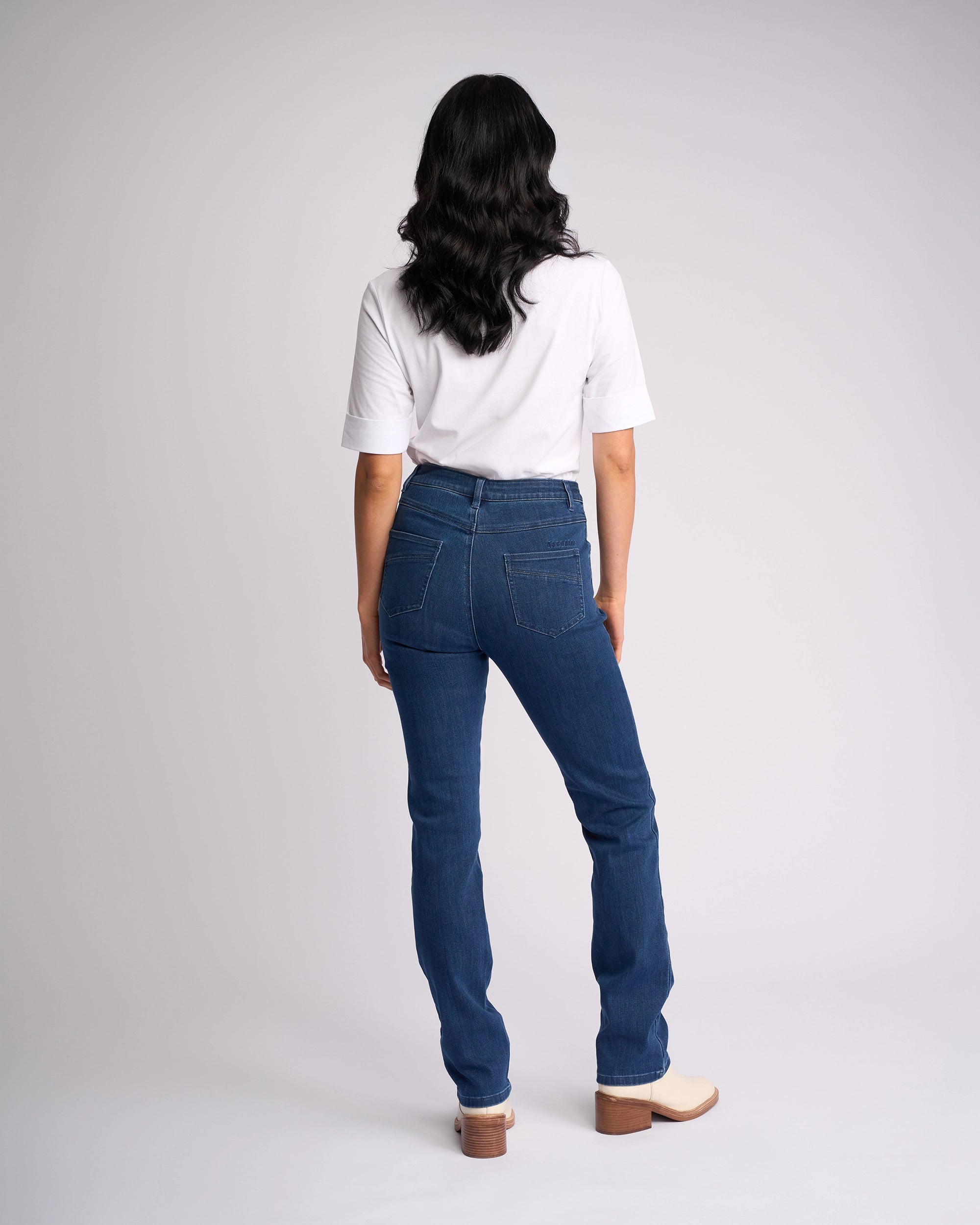A person with long, wavy dark hair is seen from behind wearing a white short-sleeve shirt and Vassalli Studio Jean - Blue Denim, paired with light heeled shoes against a plain light background.