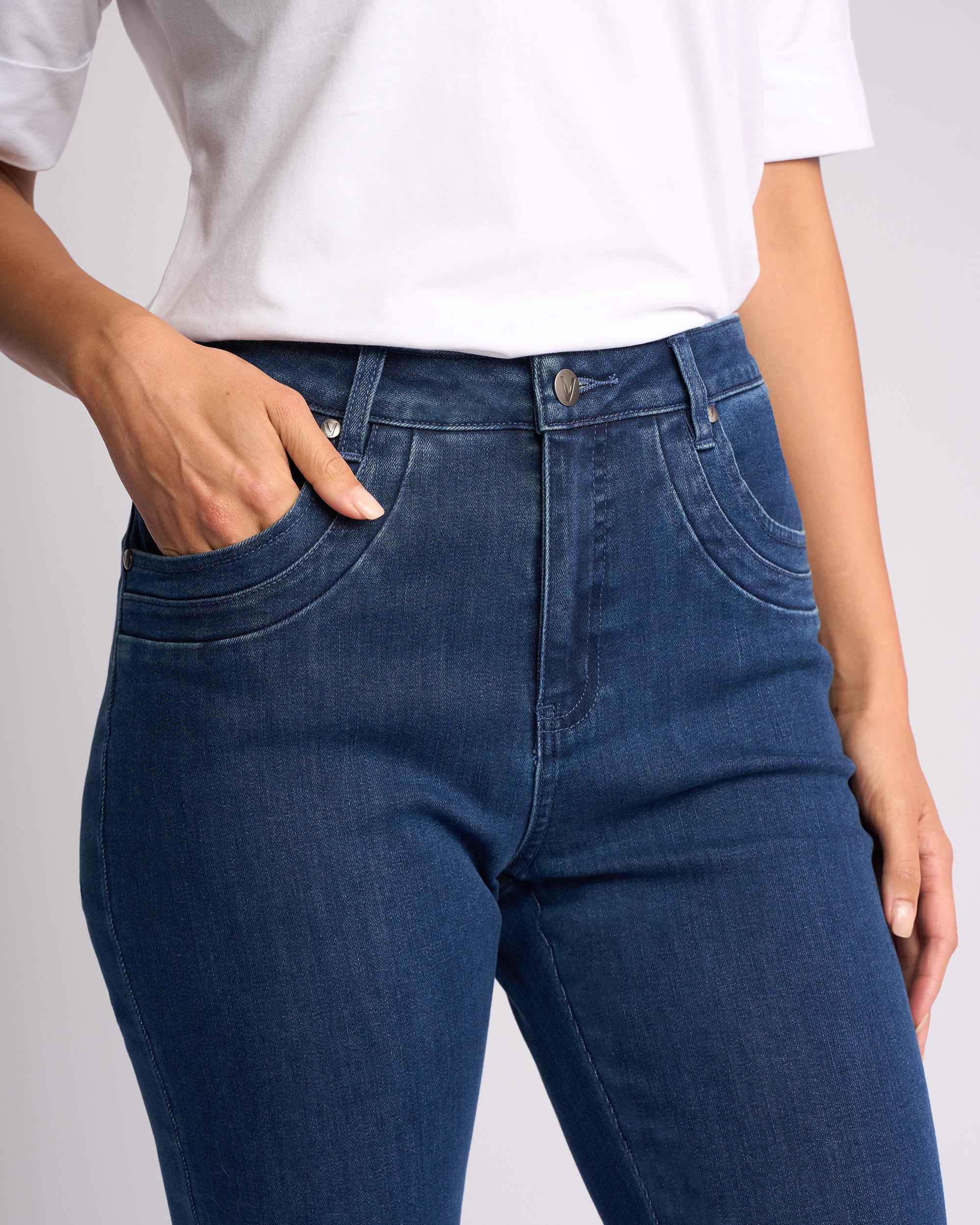 A person models the Vassalli Studio Jean - Blue Denim, pairing it with a white shirt. Shown from shoulders to thighs, they pose with one hand in a pocket and the other by their side against a plain backdrop.