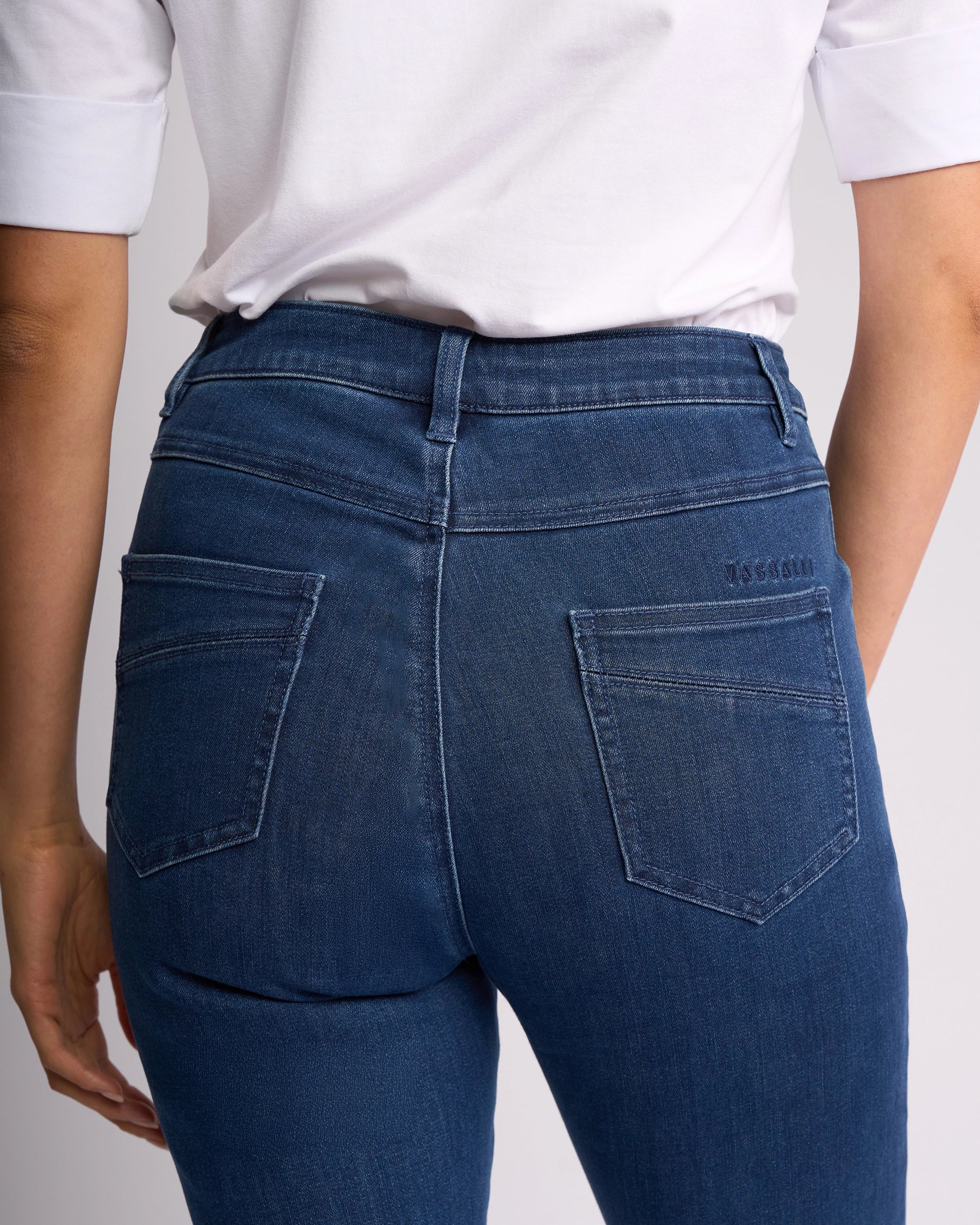 A person is shown from the back wearing Vassalli’s Studio Jean - Blue Denim, featuring two back pockets, belt loops, and a classic denim style with a white short-sleeve shirt. Focus is on the waist and upper legs.