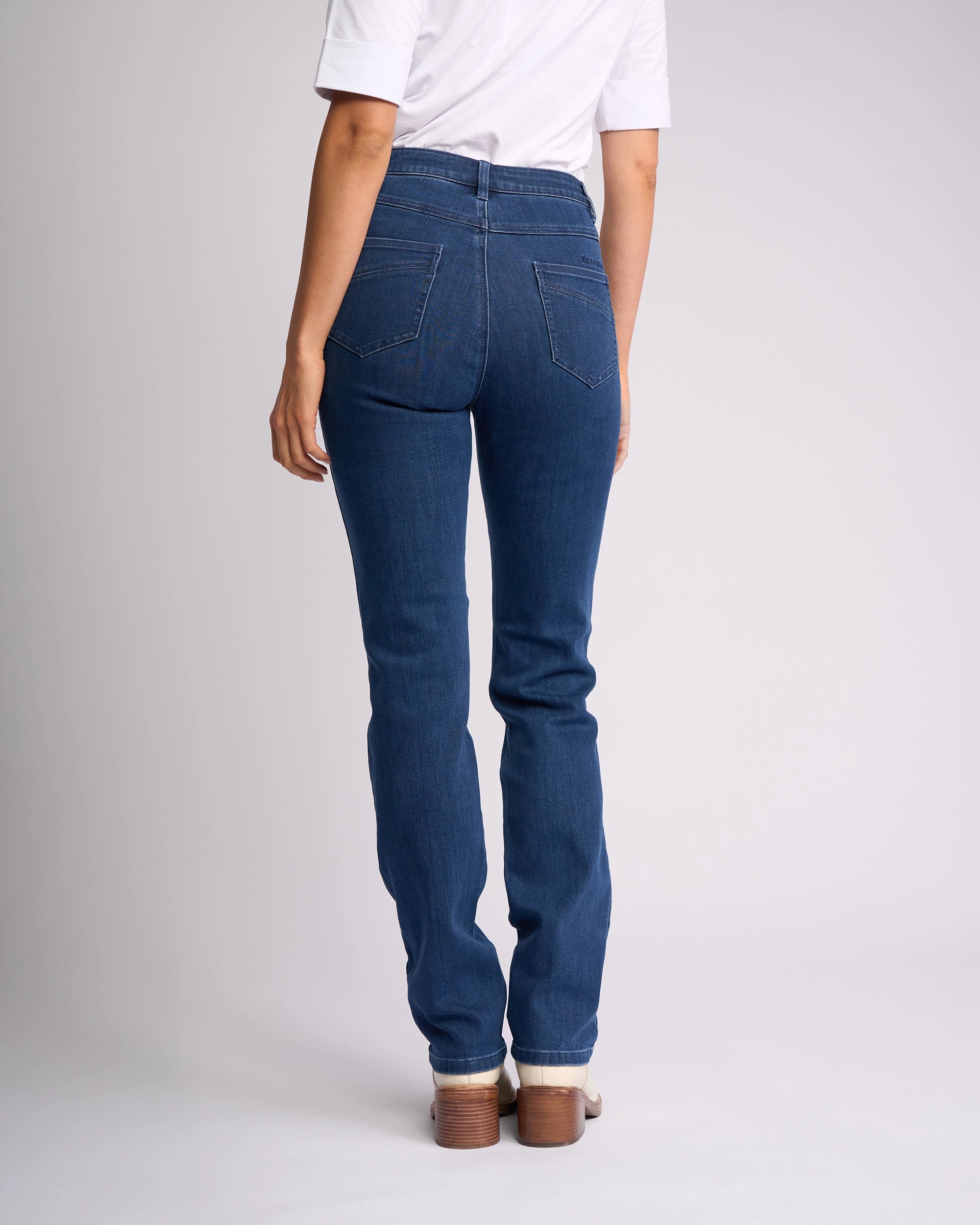 A person wearing Vassalli’s Studio Jean - Blue Denim stands facing away from the camera against a plain light background, styled with a white short-sleeve shirt and heeled shoes.