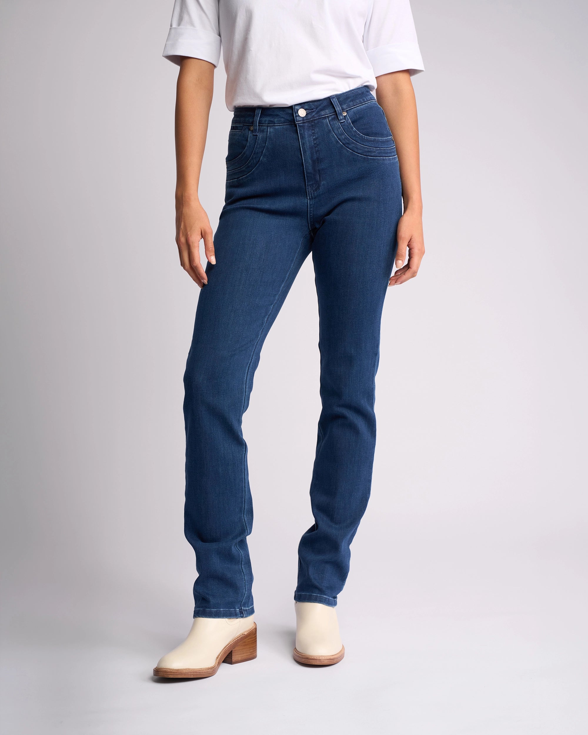A person in a white short-sleeve shirt, Vassalli Studio Jean - Blue Denim, and off-white heeled ankle boots stands against a plain light background. Their face is not visible.