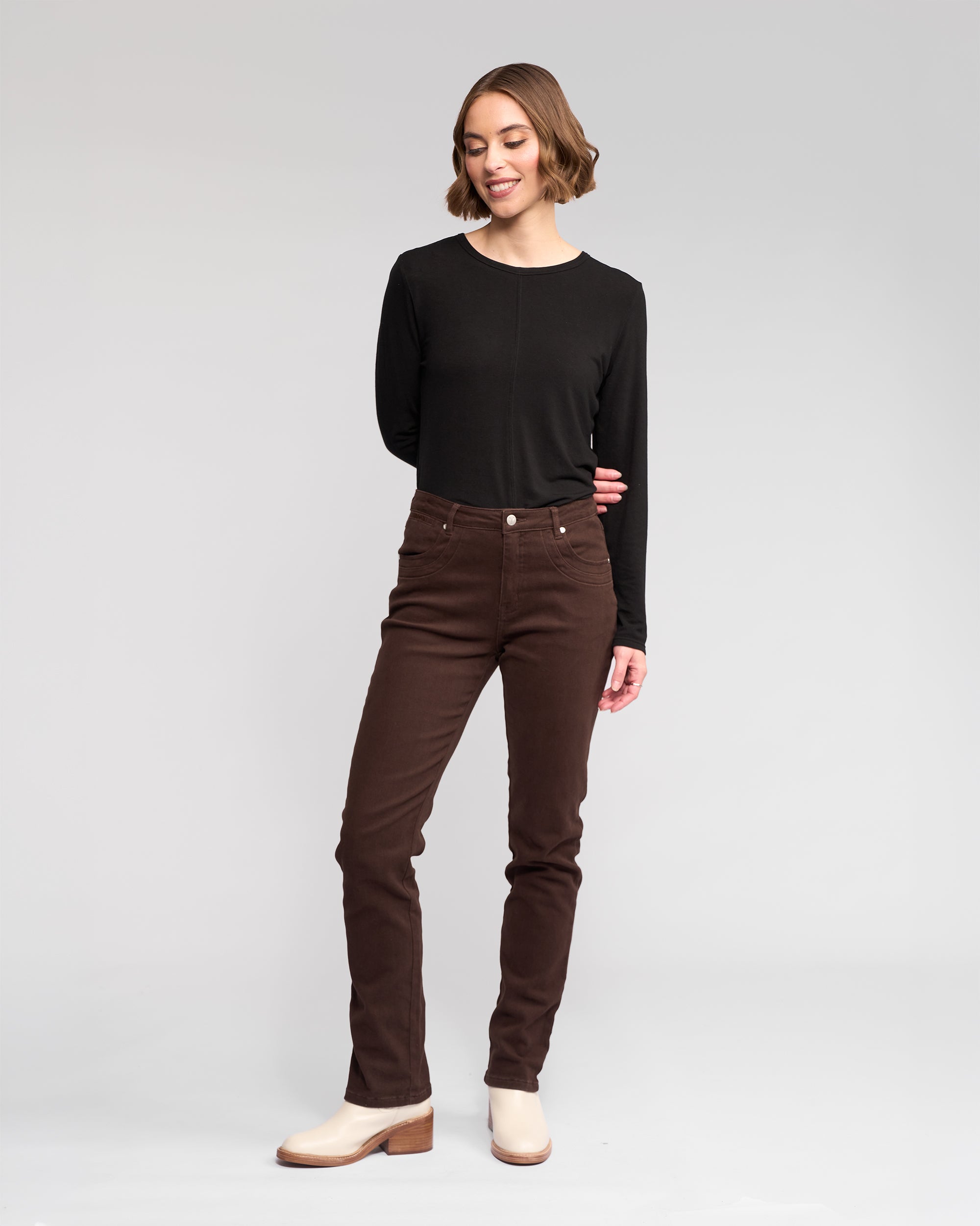 A woman stands against a plain light background, smiling in a Vassalli Studio Jean - Chocolate and a long-sleeve black top with light-colored ankle boots, her arms relaxed behind her back as she looks slightly to the side.