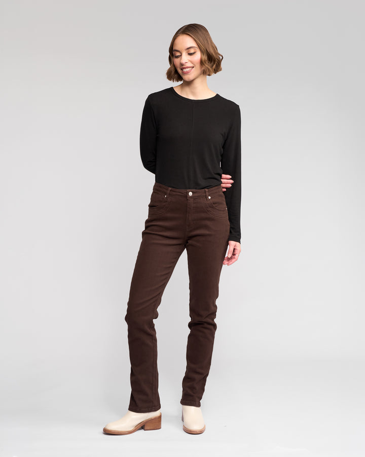 A woman stands against a plain light background, smiling in a Vassalli Studio Jean - Chocolate and a long-sleeve black top with light-colored ankle boots, her arms relaxed behind her back as she looks slightly to the side.