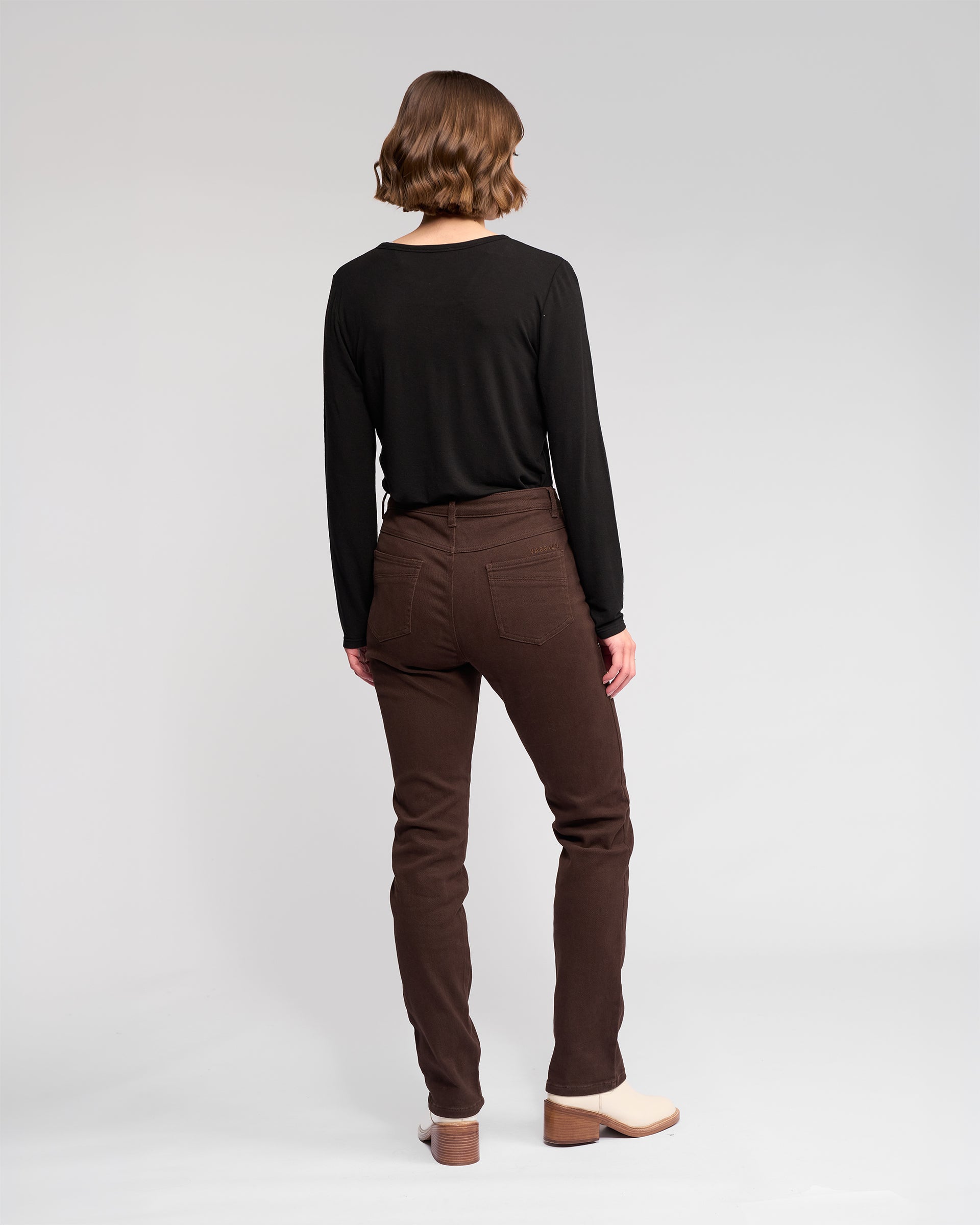 A person with short, wavy brown hair stands with their back to the camera, wearing a long-sleeved black shirt and Vassalli Studio Jean - Chocolate, paired with cream-colored heeled shoes against a plain light background.