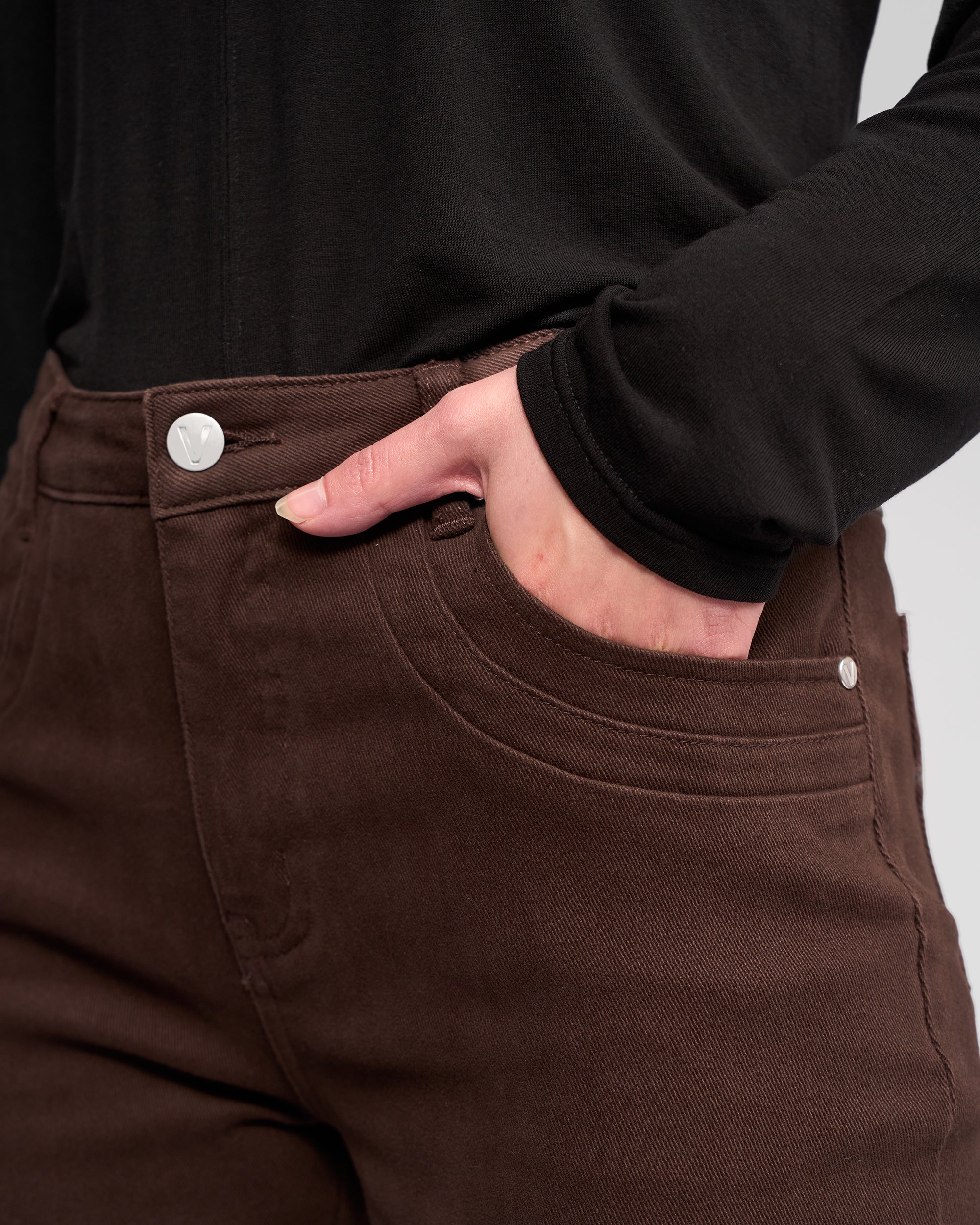 A person wearing a black long-sleeve top and Vassalli Studio Jean - Chocolate, with their right hand in the front pocket of the jeans.