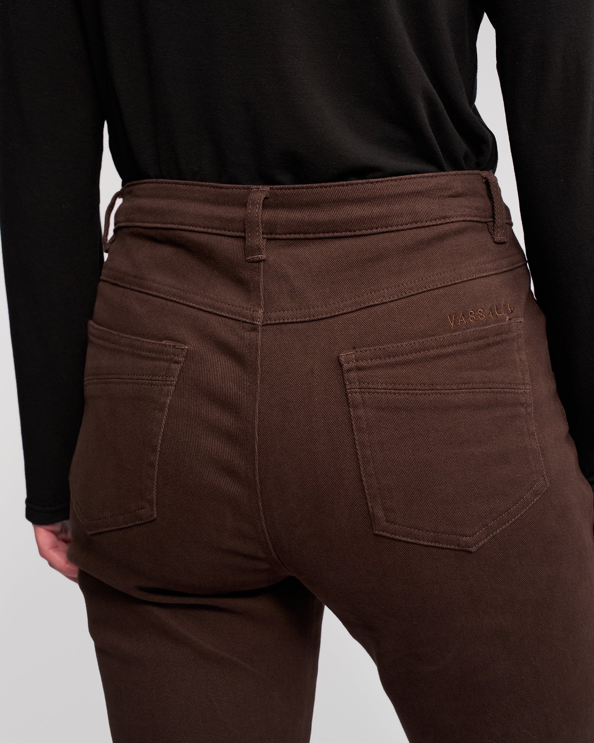 A person is shown from the back wearing Vassalli Studio Jean in chocolate, a stretch denim slim-leg style with two back pockets and a small embroidered logo near the waistband, paired with a black long-sleeve top.