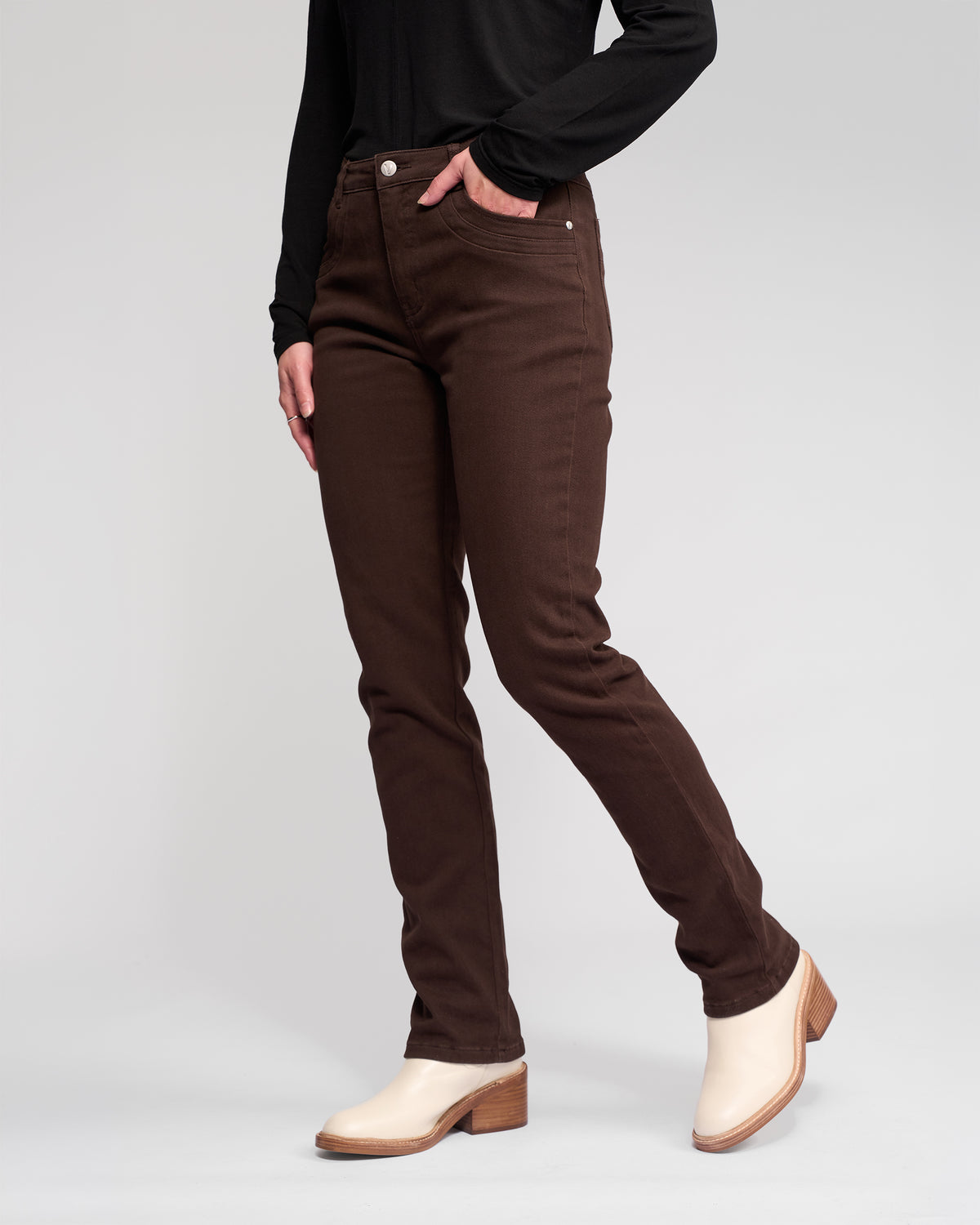 A person stands against a plain light gray background wearing Vassalli Studio Jean - Chocolate, a black long-sleeve top, and off-white heeled ankle boots. The upper body is partially out of frame.