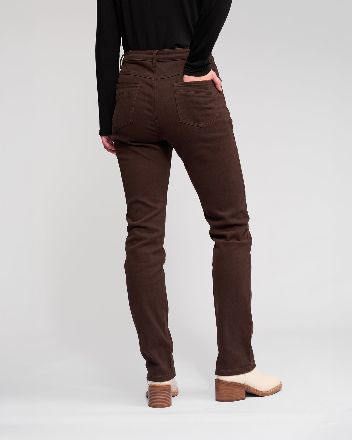 A person stands facing away in Vassalli's Studio Jean - Chocolate, high-waisted stretch denim slim-leg jeans, paired with a black long-sleeve top and beige heeled boots, one hand in a back pocket, against a plain light background.