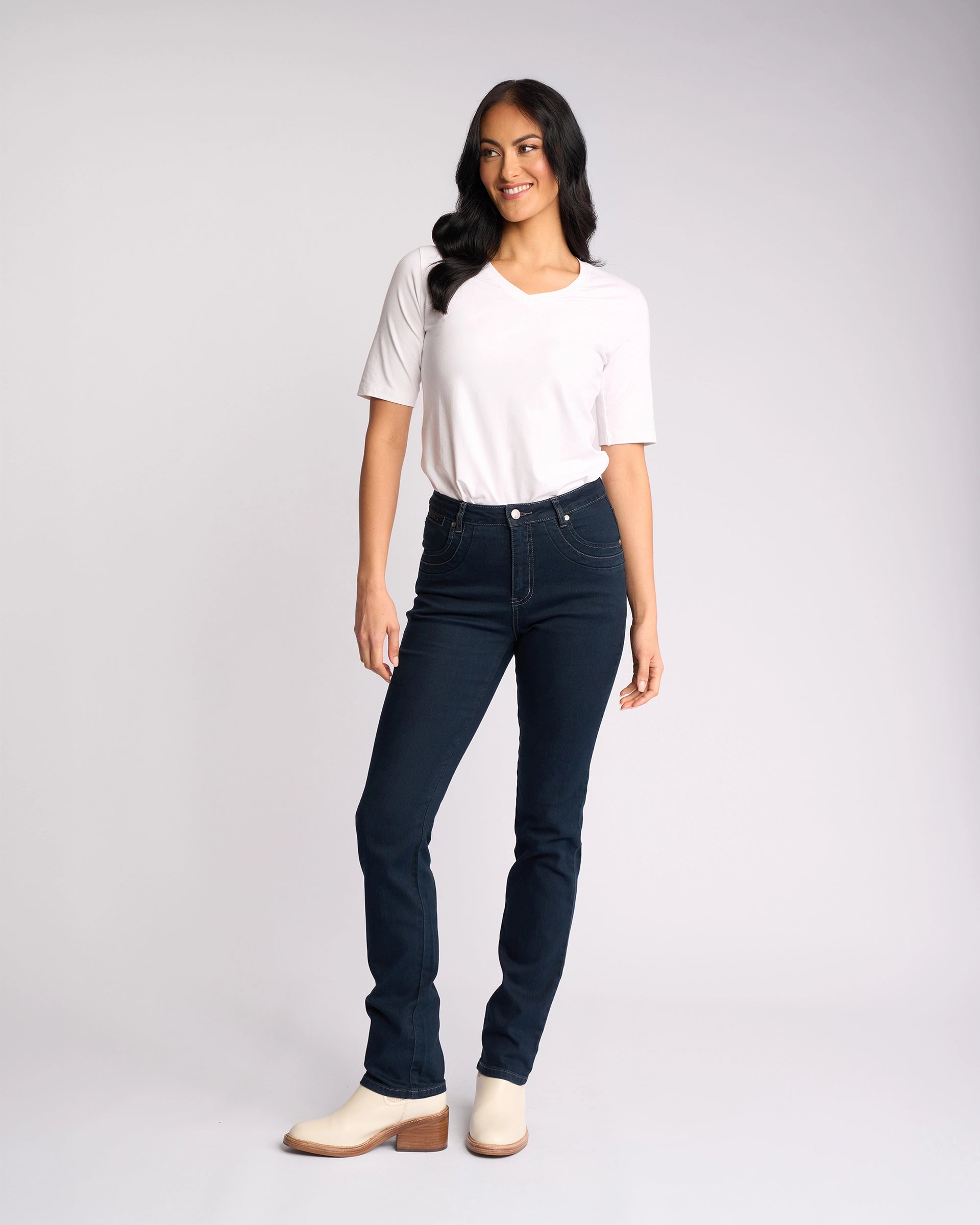 A woman, smiling and looking to the side, stands against a plain light background wearing Vassalli’s Studio Jean in Indigo Denim with a white short-sleeve shirt and light heeled boots. One hand is relaxed by her side.