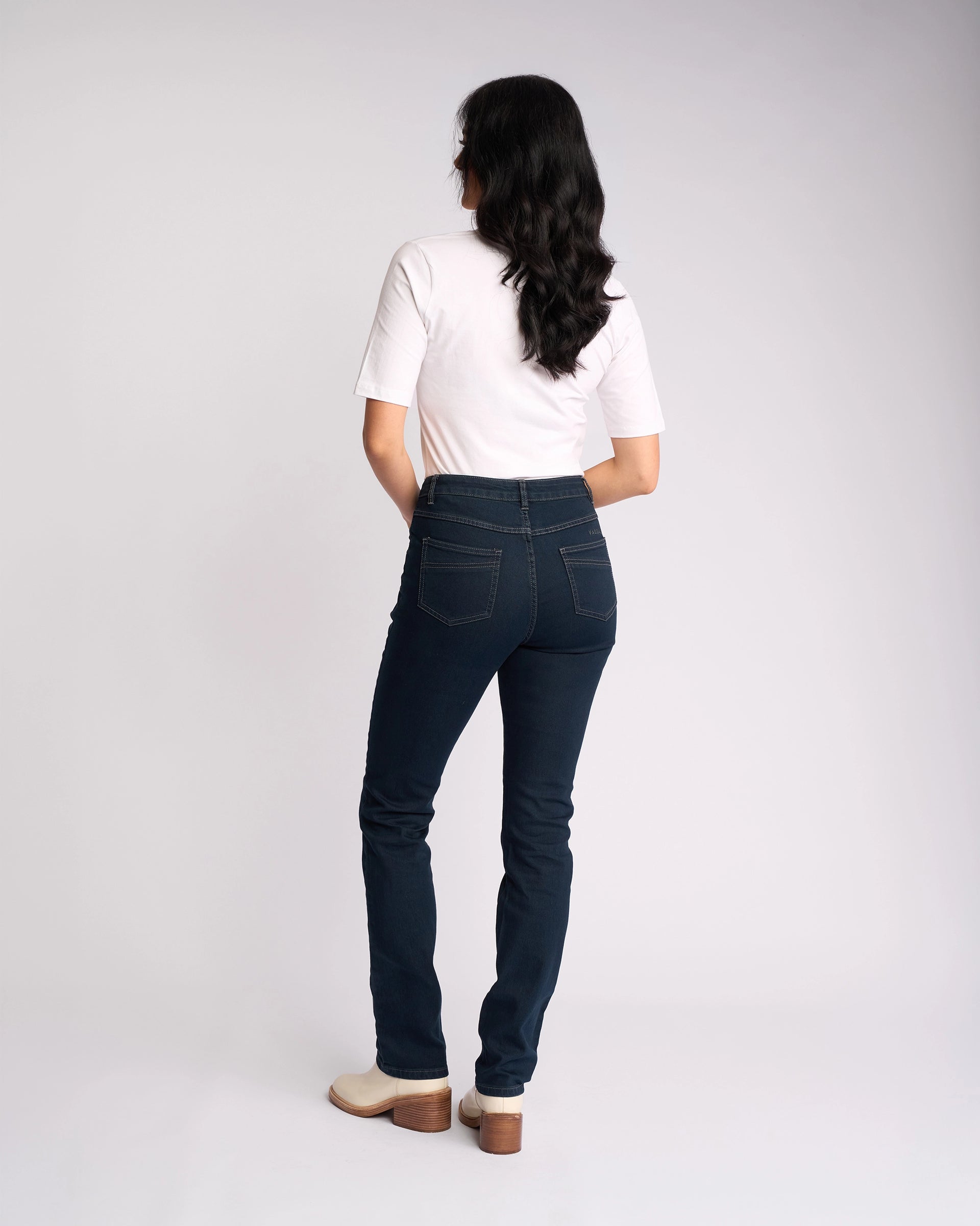 A person with long dark hair, wearing a white short-sleeve shirt, Vassalli Studio Jean in Indigo Denim, and beige heeled shoes stands facing away against a plain light background.