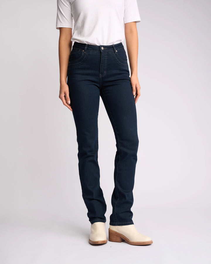 A person wearing a white short-sleeve top, Vassalli Studio Jean - Indigo Denim, and light heeled ankle boots stands against a plain light background. The person’s face is not visible.