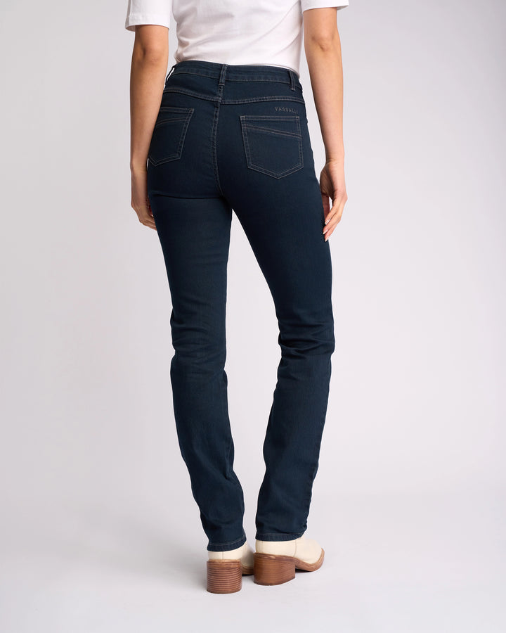 Someone stands with their back to the camera against a plain light background, wearing Vassalli’s Studio Jean in Indigo Denim, paired with a white top and light-colored heeled shoes.