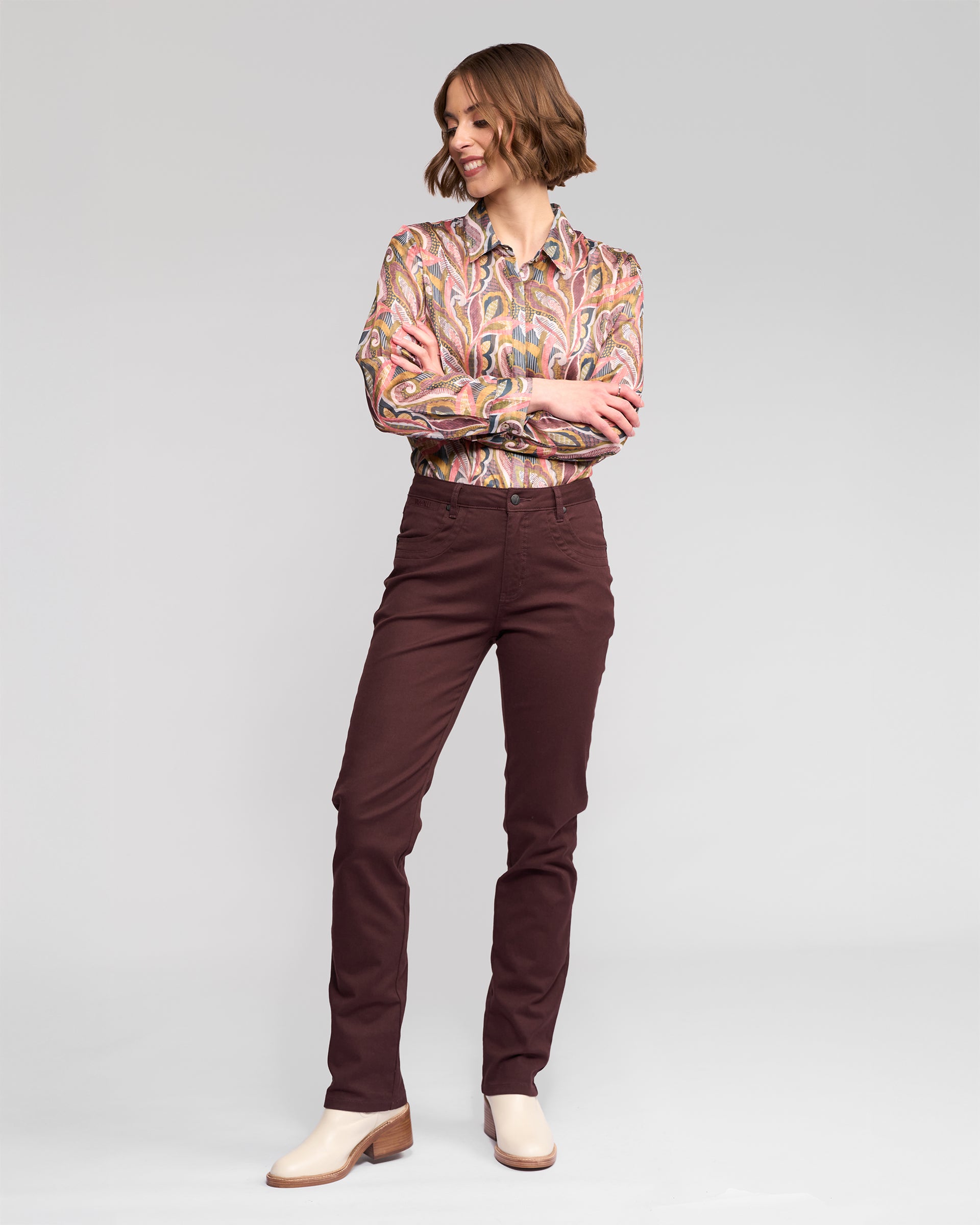 A woman with short wavy hair smiles, wearing a colorful long-sleeve blouse, Vassalli Studio Jean - Merlot slim-leg jeans, and cream ankle boots against a plain light background.