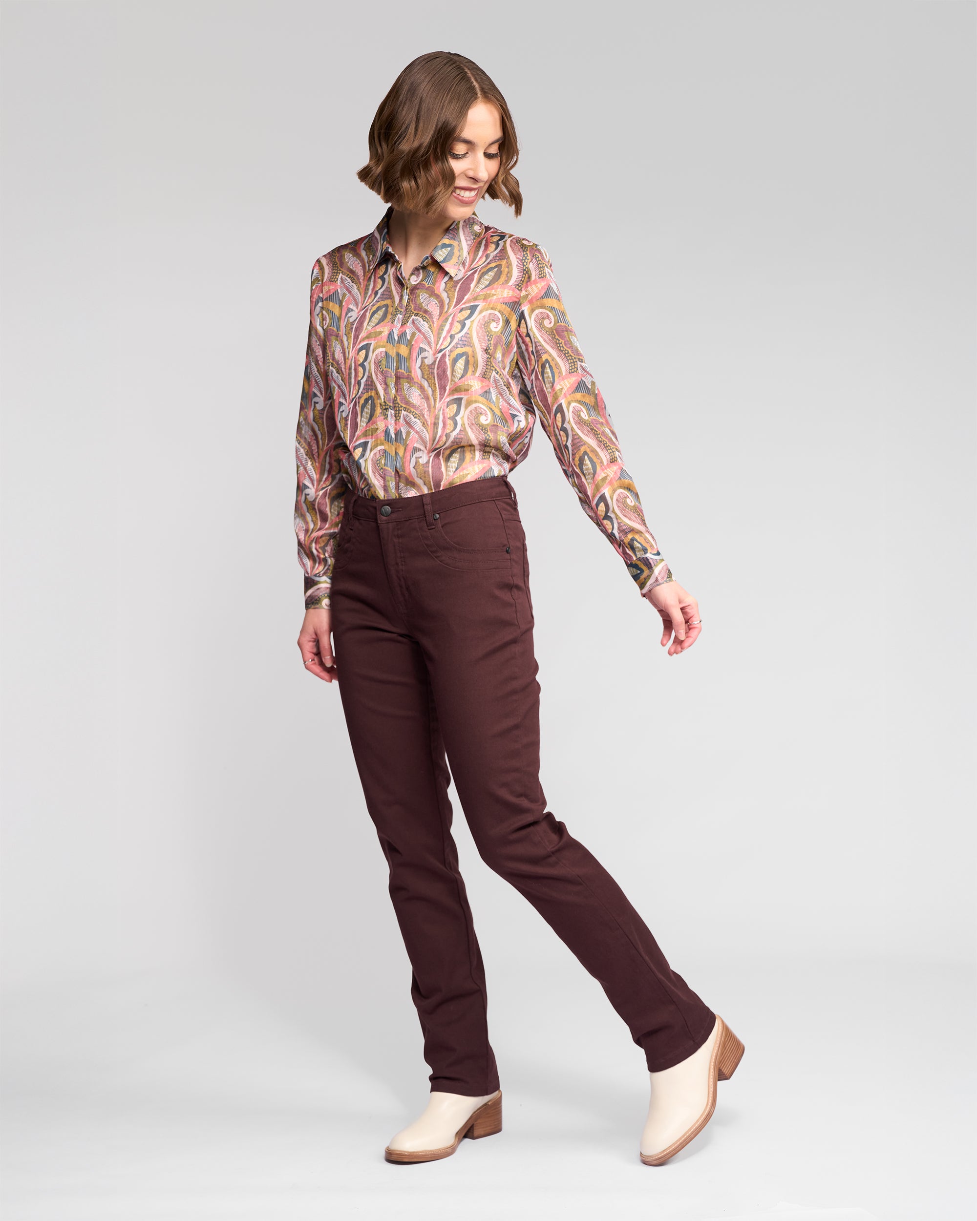 A woman poses in a studio wearing Vassalli's Studio Jean - Merlot, paired with a patterned long-sleeve shirt and cream ankle boots. She looks down and slightly to the side, one leg extended.