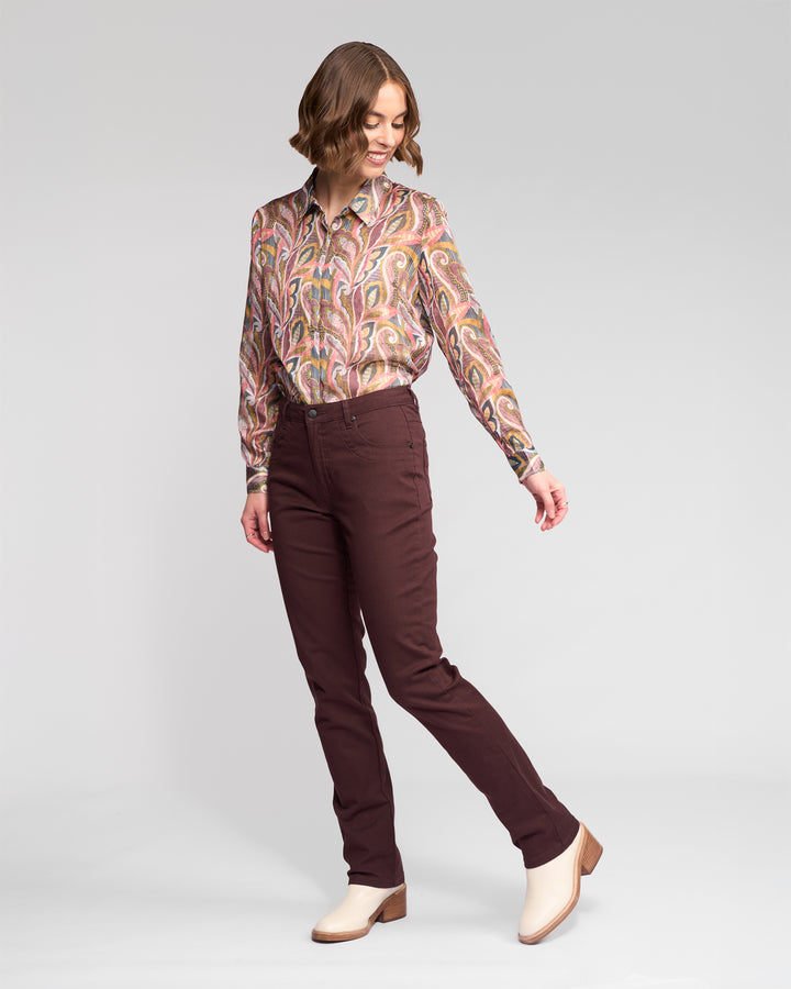A woman poses in a studio wearing Vassalli's Studio Jean - Merlot, paired with a patterned long-sleeve shirt and cream ankle boots. She looks down and slightly to the side, one leg extended.