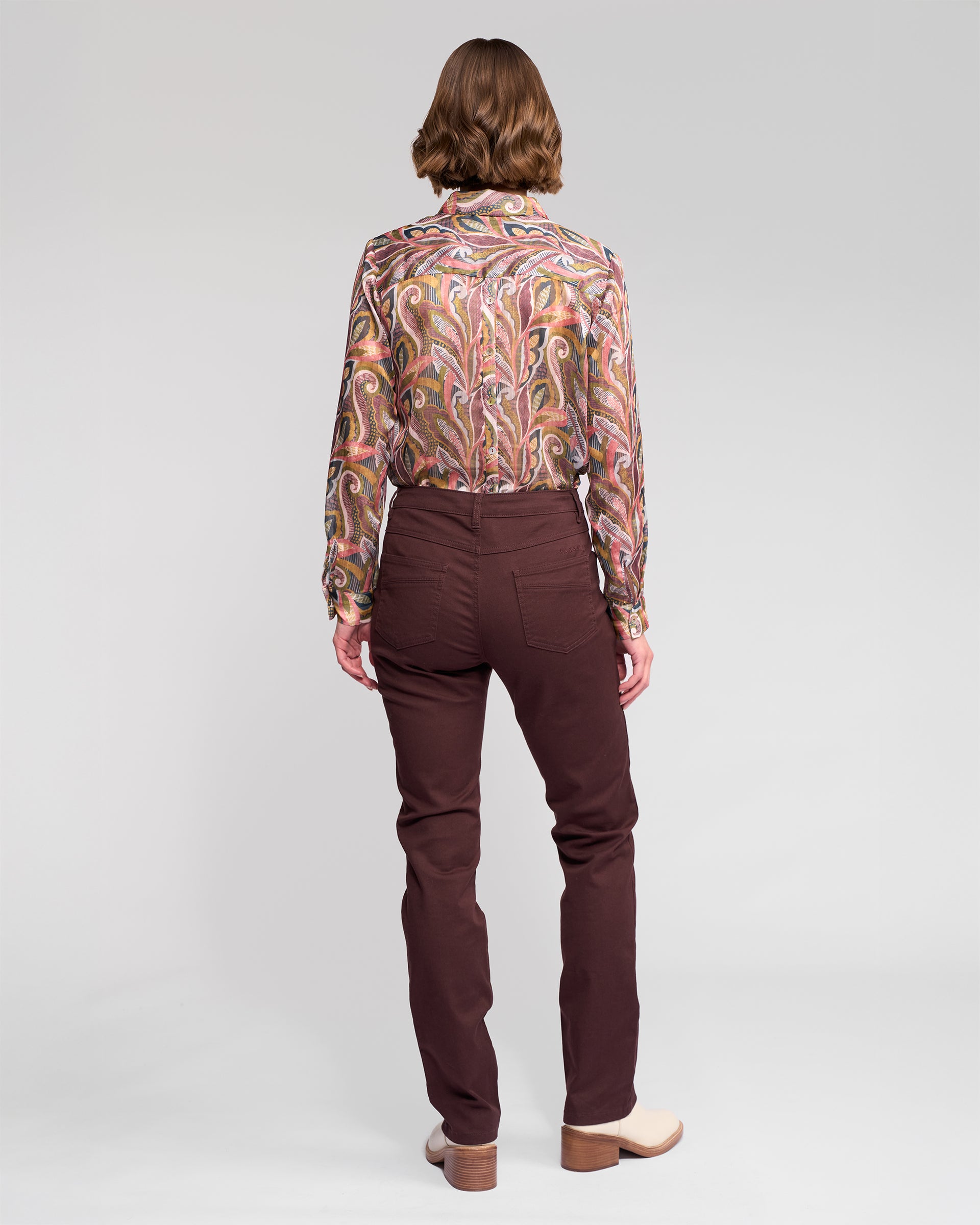 A person with short brown hair stands facing away, wearing a colorful paisley blouse, Vassalli Studio Jean - Merlot, and light shoes against a plain light background.