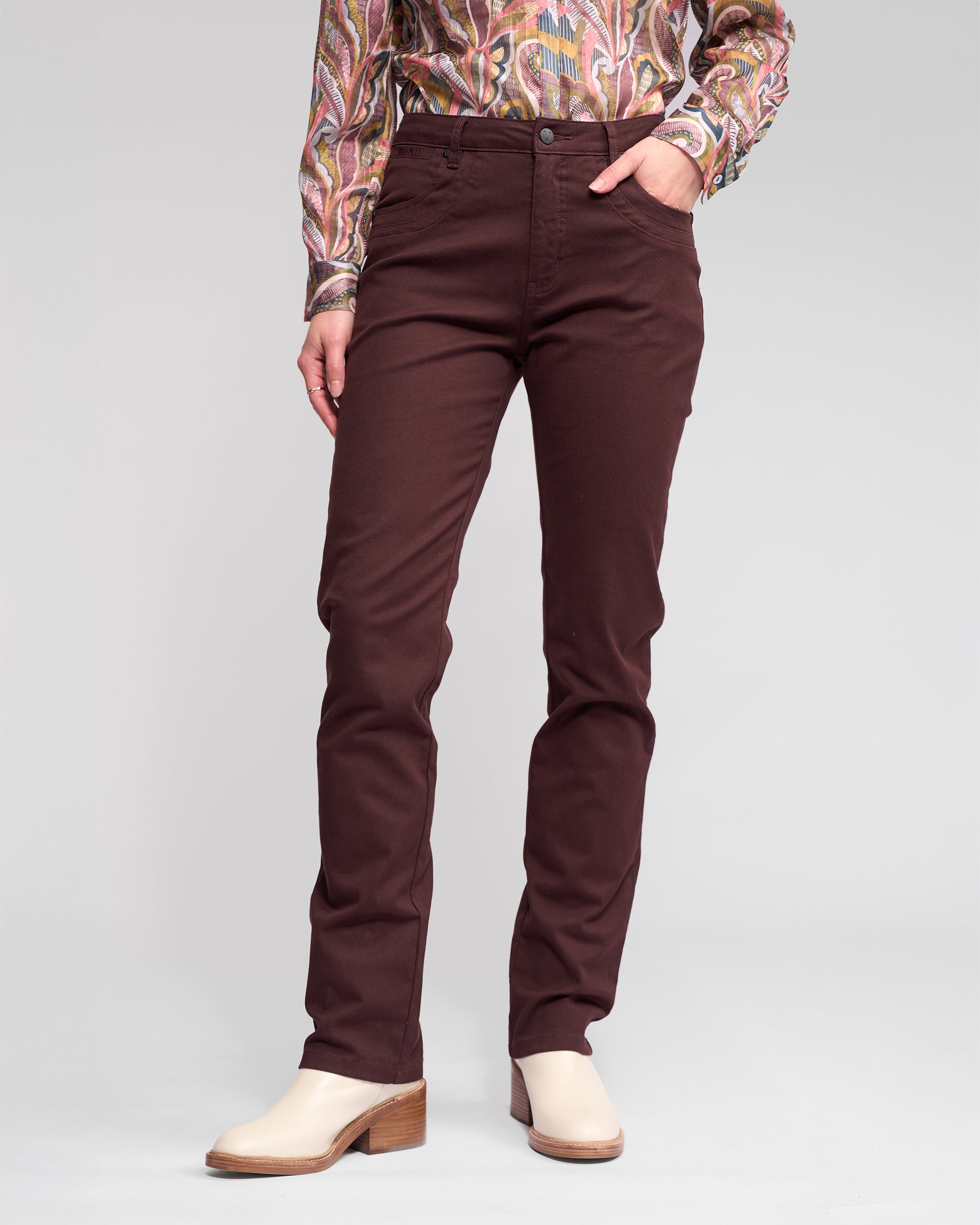 Someone wearing Vassalli's Studio Jean in Merlot, a patterned long-sleeve shirt, and cream heeled shoes stands against a plain light gray background with one hand in their pocket.
