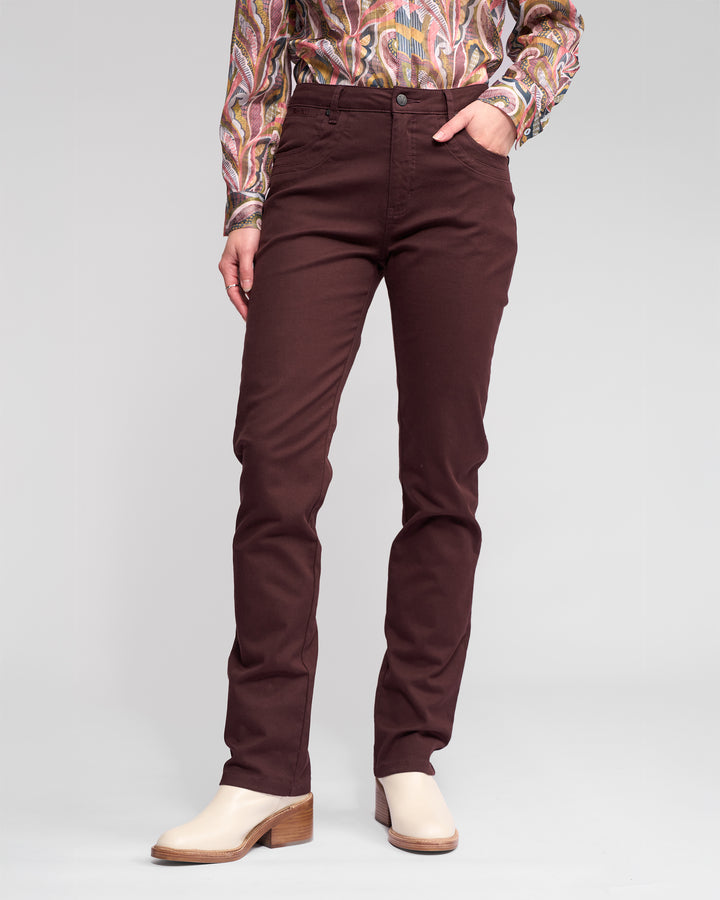 Someone wearing Vassalli's Studio Jean in Merlot, a patterned long-sleeve shirt, and cream heeled shoes stands against a plain light gray background with one hand in their pocket.