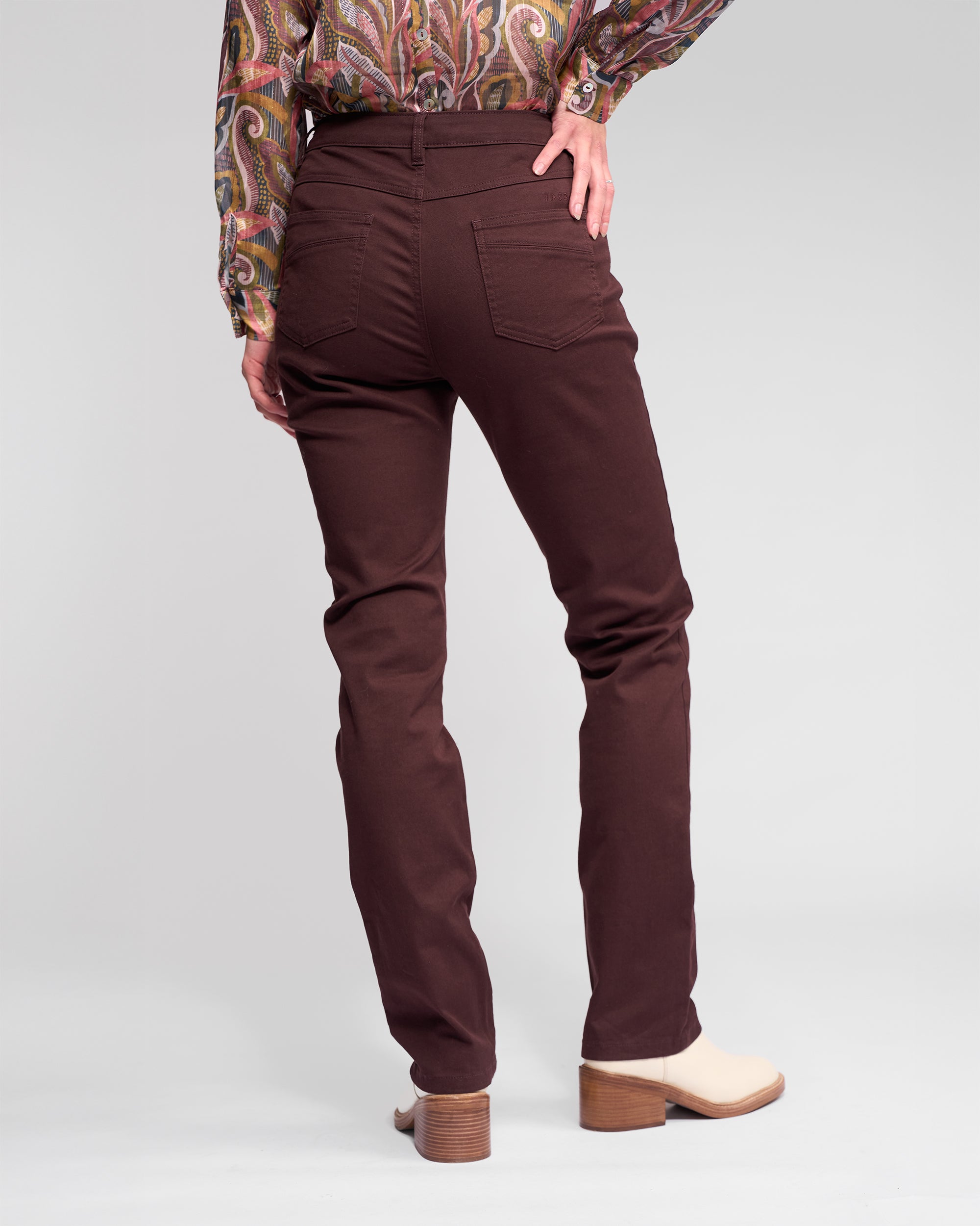 Person stands with their back to the camera, wearing Vassalli's Studio Jean in Merlot, a paisley shirt, and beige heeled ankle boots against a plain light background.