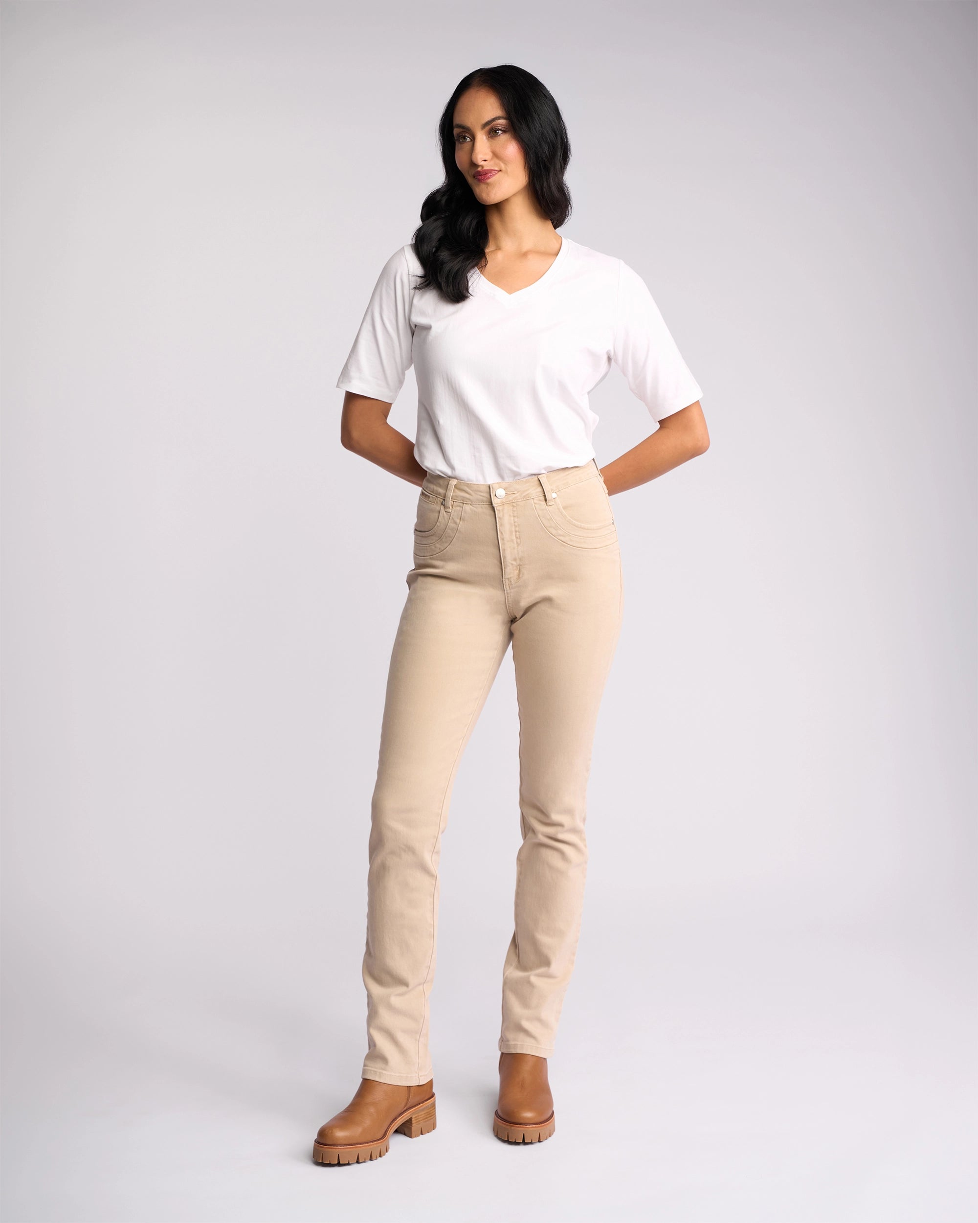 A woman wearing Vassalli’s Studio Jean in Oat Denim stands against a plain light background, paired with a white V-neck tee and brown boots, arms behind her back as she gazes slightly to the side.