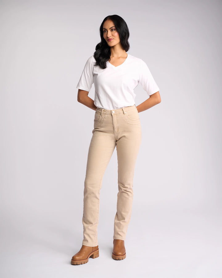 A woman wearing Vassalli’s Studio Jean in Oat Denim stands against a plain light background, paired with a white V-neck tee and brown boots, arms behind her back as she gazes slightly to the side.