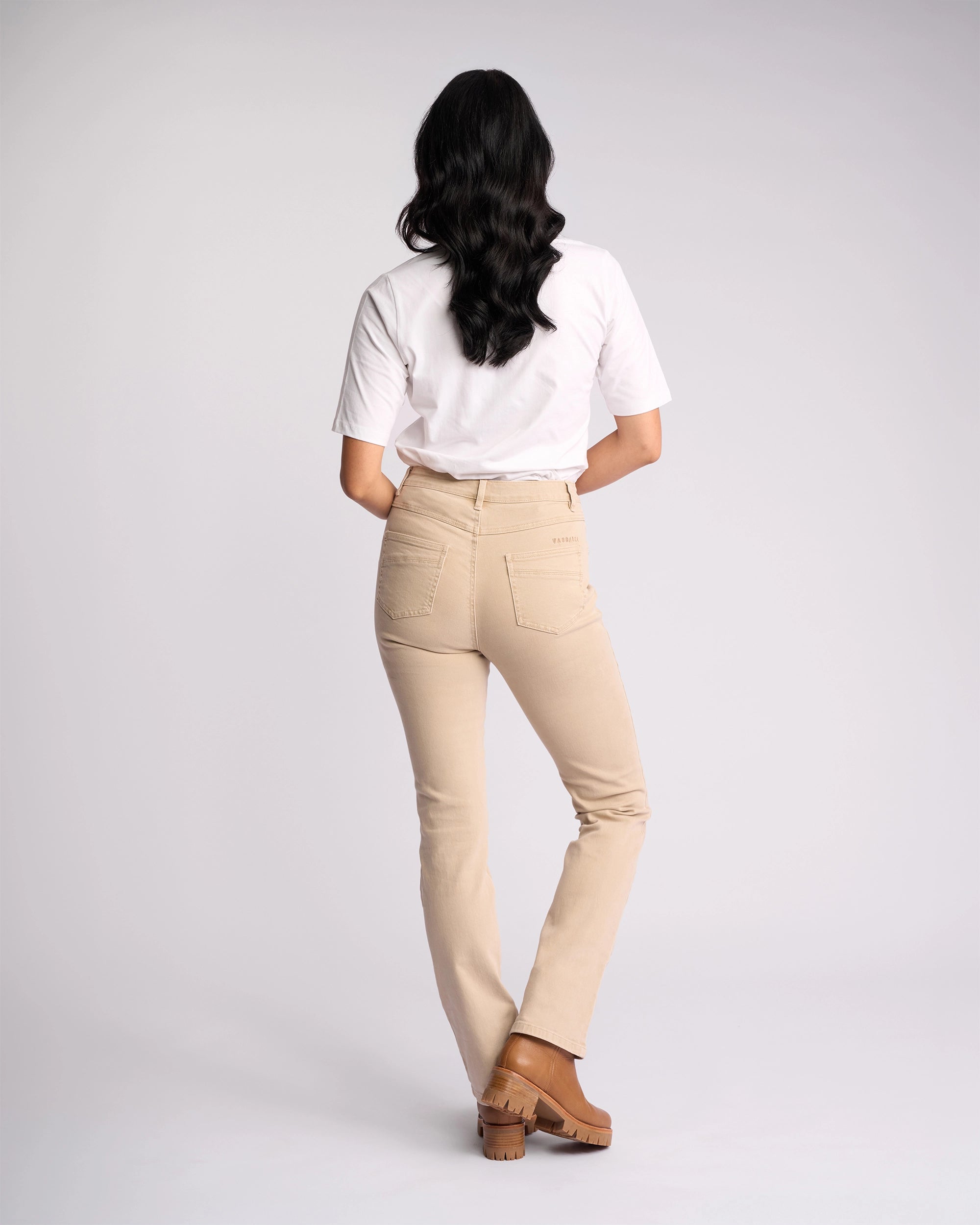 A person with long dark hair, wearing a white t-shirt, Vassalli Studio Jean in Oat Denim, and brown shoes stands facing away against a plain light background.