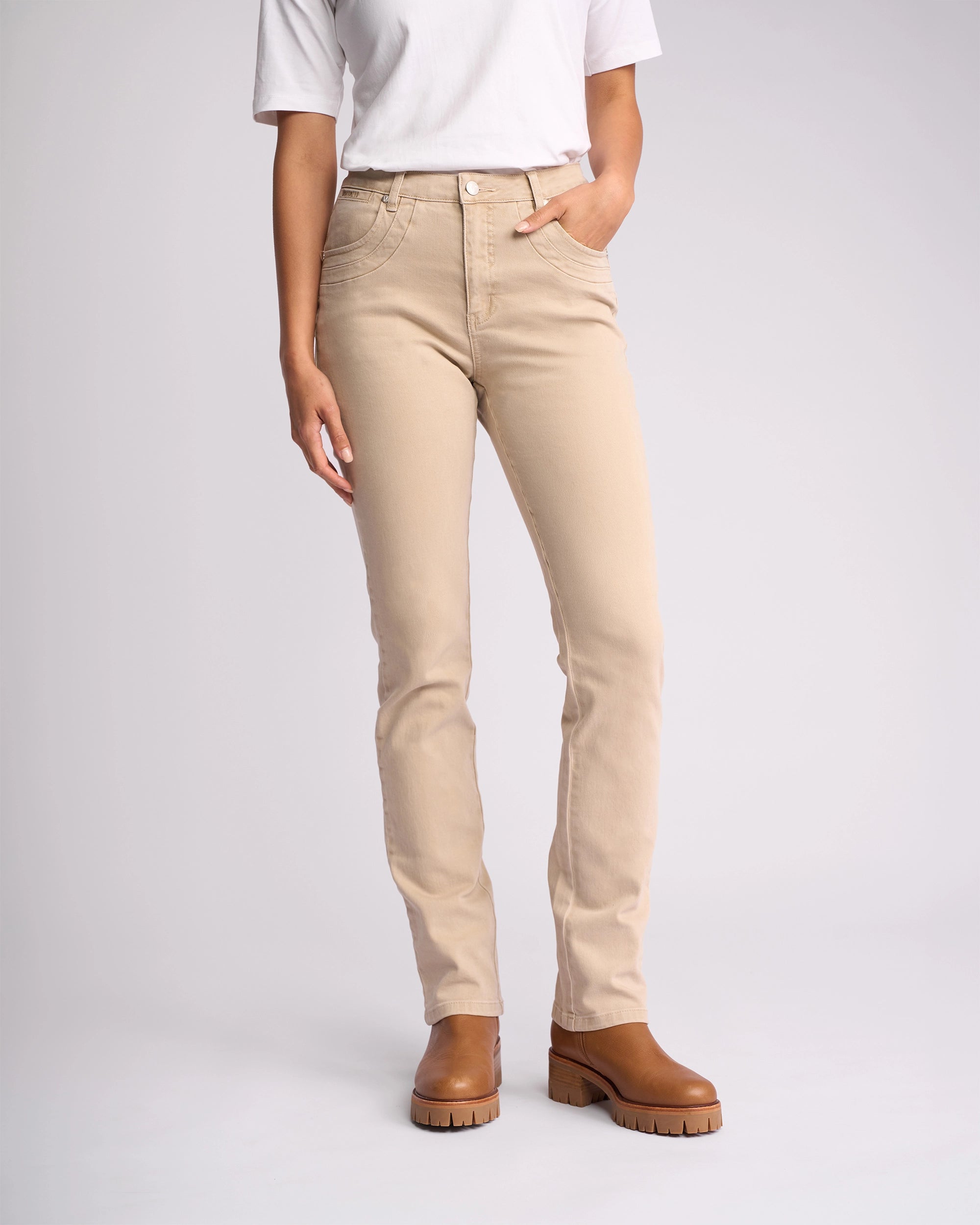 A person stands against a plain light background wearing a white short-sleeve shirt, Vassalli Studio Jean - Oat Denim, and brown ankle boots. Their left hand is in their pocket, and their face isn't visible.