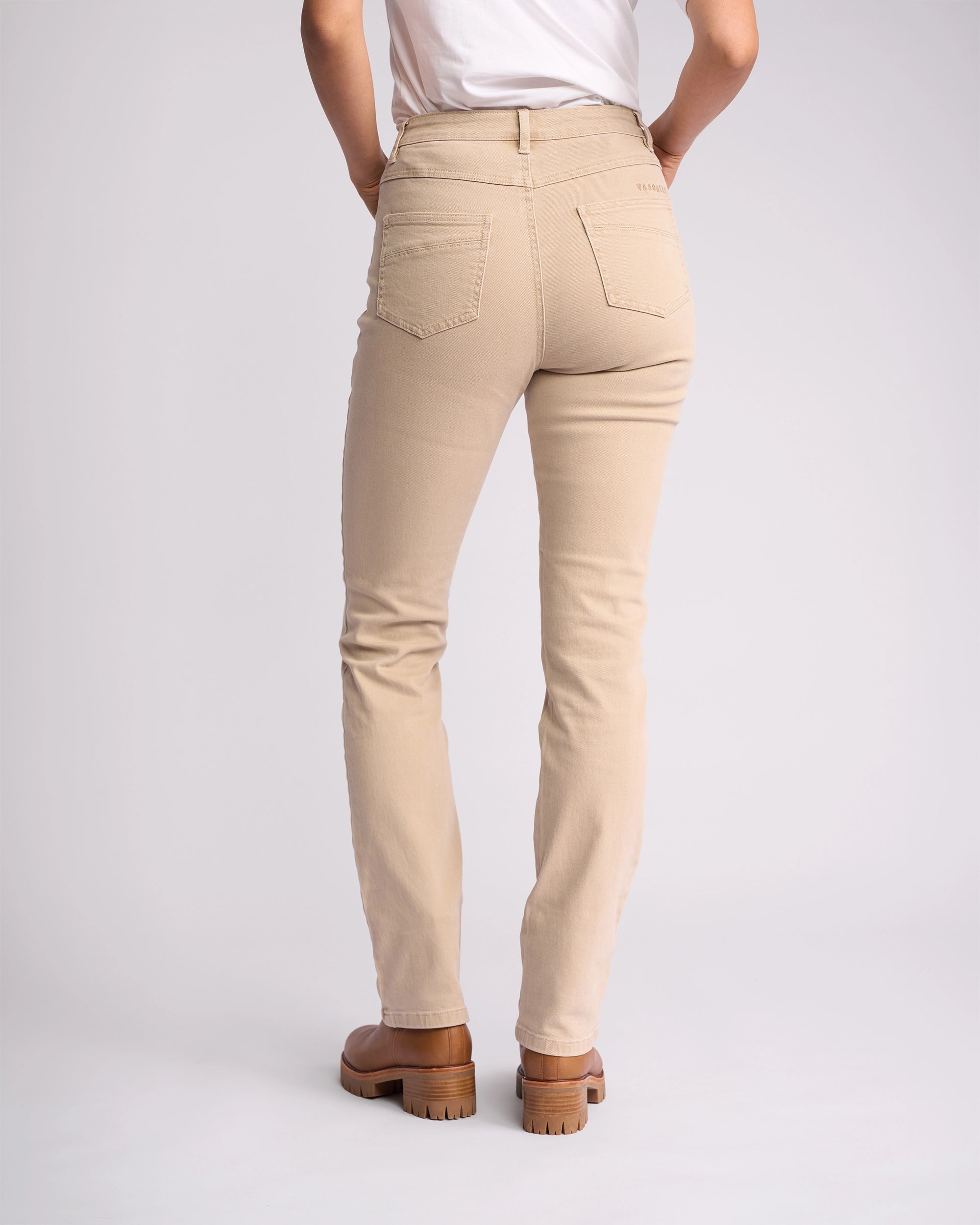A person is standing with their back to the camera against a plain light background, wearing Vassalli’s Studio Jean in Oat Denim, styled with a white top and brown heeled shoes.