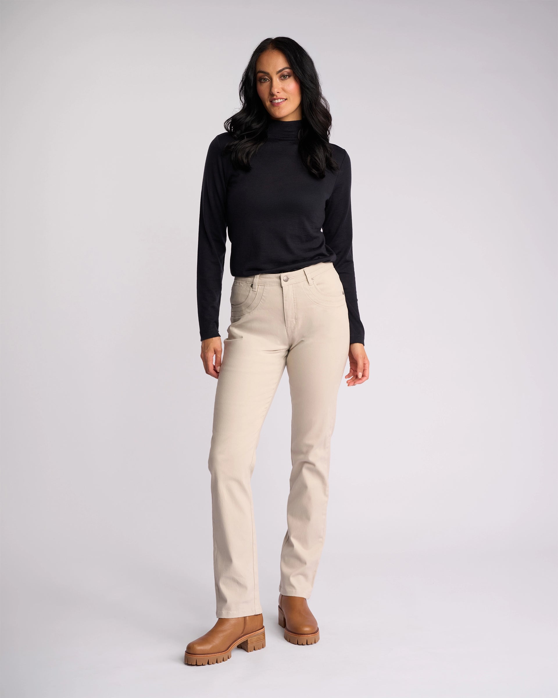 A woman with long dark hair wears Vassalli's Studio Jean in Pumice, a black long-sleeve top, and brown boots, standing against a plain light gray background.