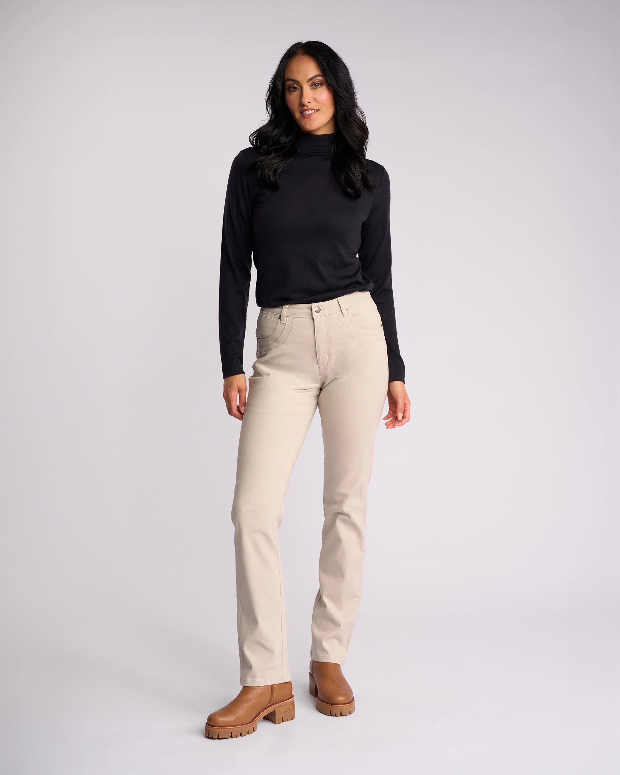 A woman with long dark hair wears Vassalli's Studio Jean in Pumice, a black long-sleeve top, and brown boots, standing against a plain light gray background.