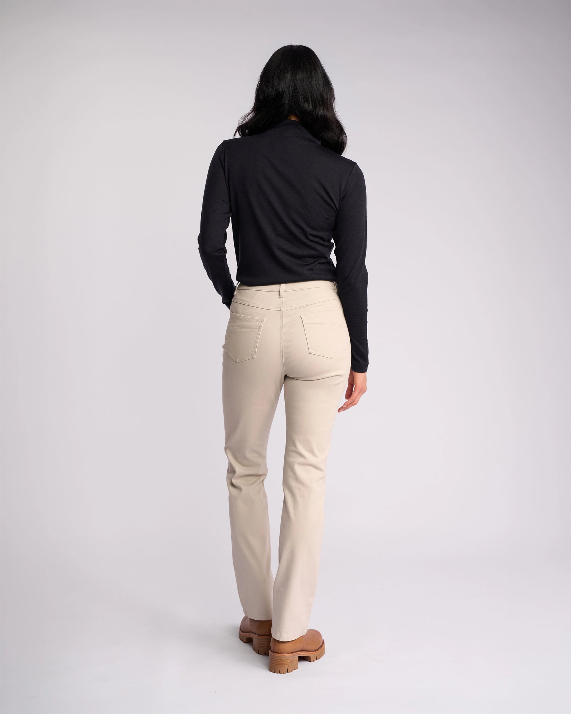 A person with long dark hair, wearing a black long-sleeve top and Vassalli Studio Jean - Pumice, stands facing away from the camera against a plain light background.