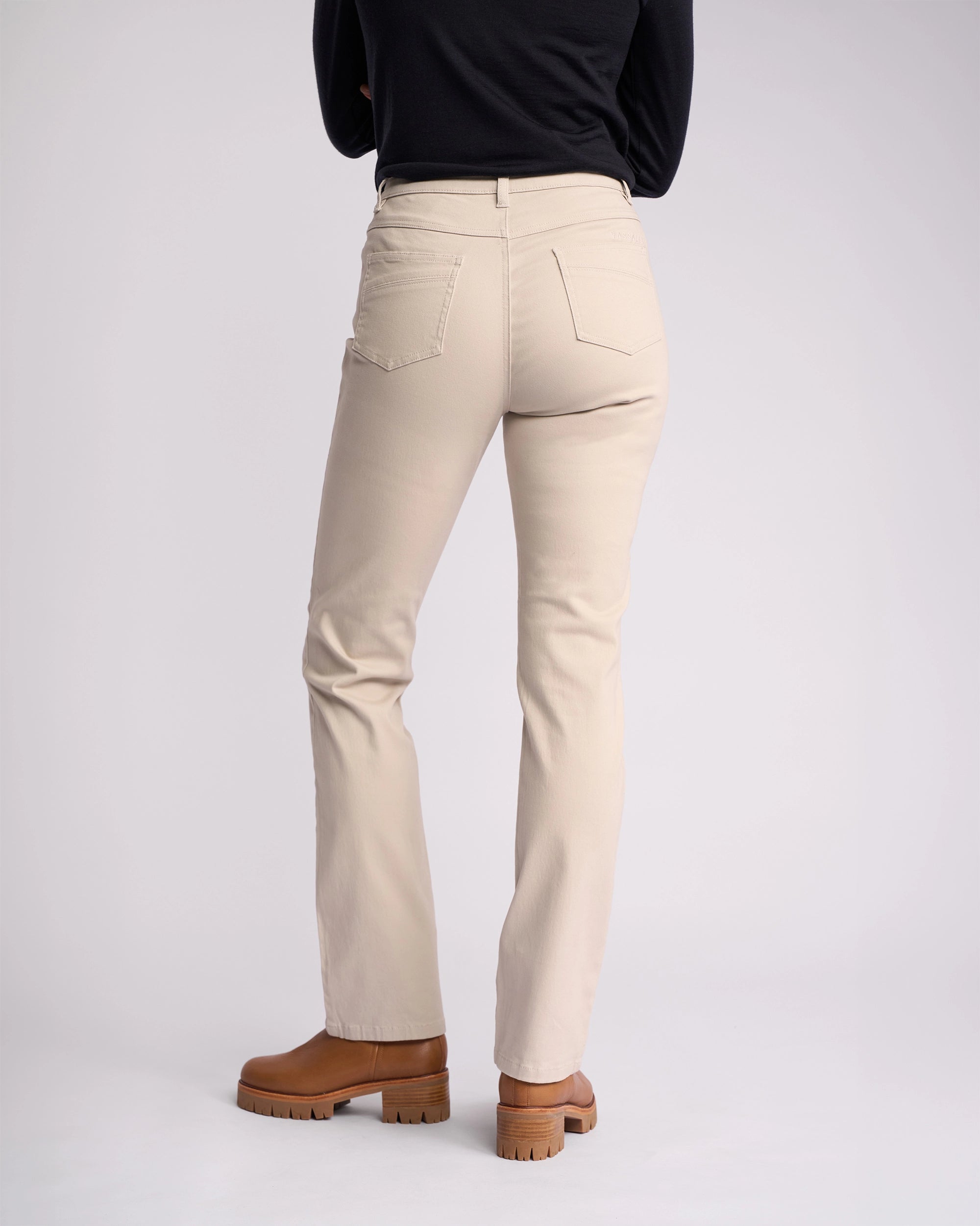 A person wearing Vassalli Studio Jean - Pumice stands facing away from the camera in stretch denim, paired with a black long-sleeve top and brown boots against a plain light background.