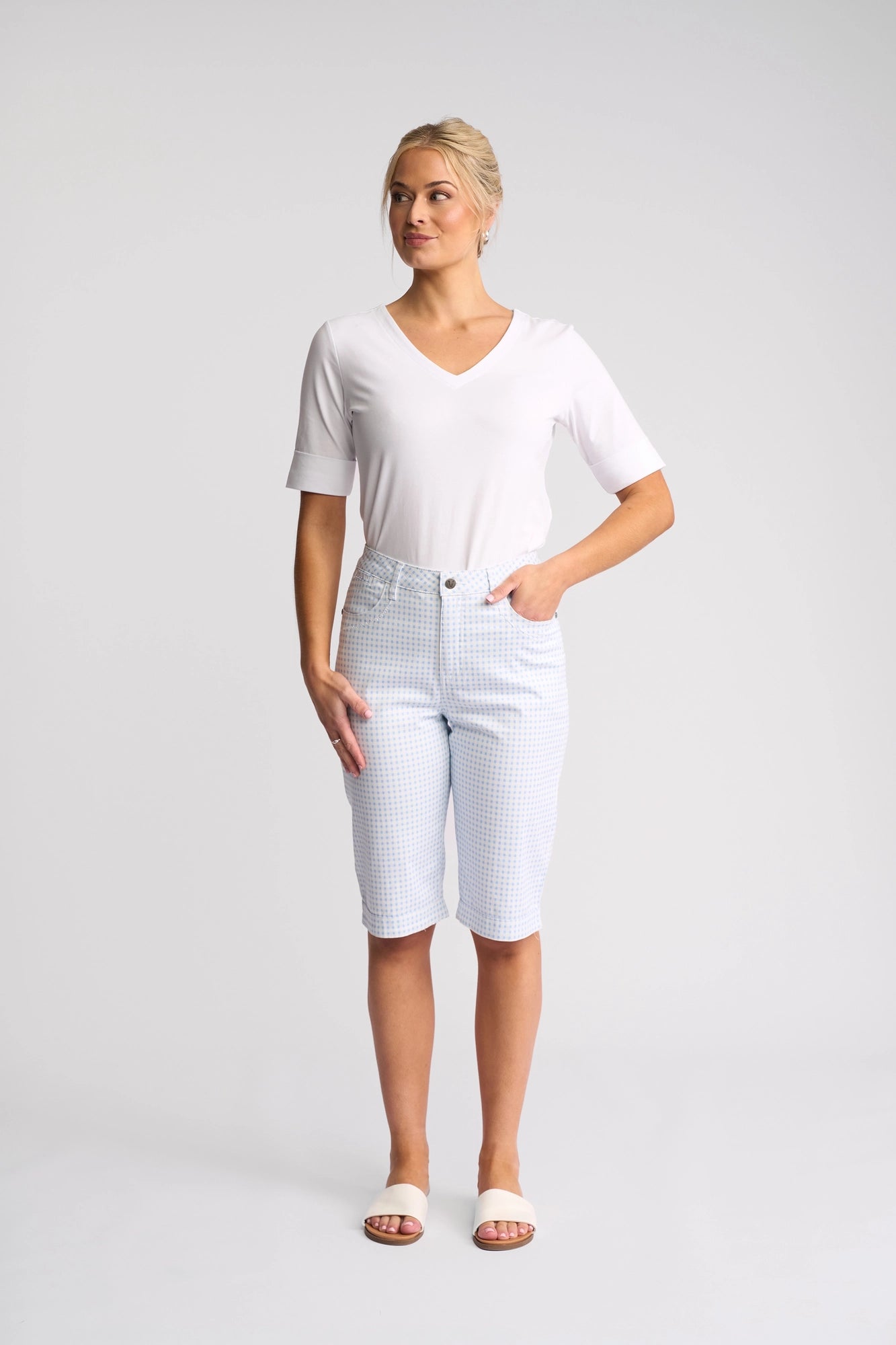 A woman stands against a plain background wearing a white V-neck top, white sandals, and the Vassalli Custom Short in Blue Gingham. With one hand in her pocket and gazing off to the side, she effortlessly highlights the relaxed style of these lightweight shorts.