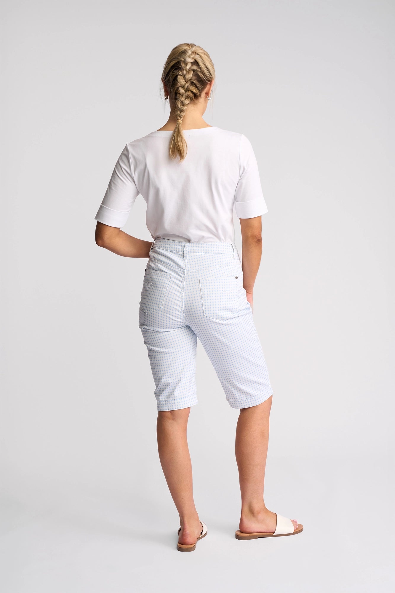 A woman with blonde braided hair, wearing a white T-shirt, Vassalli’s Custom Short in Blue Gingham, and white slide sandals stands facing away against a plain white background.
