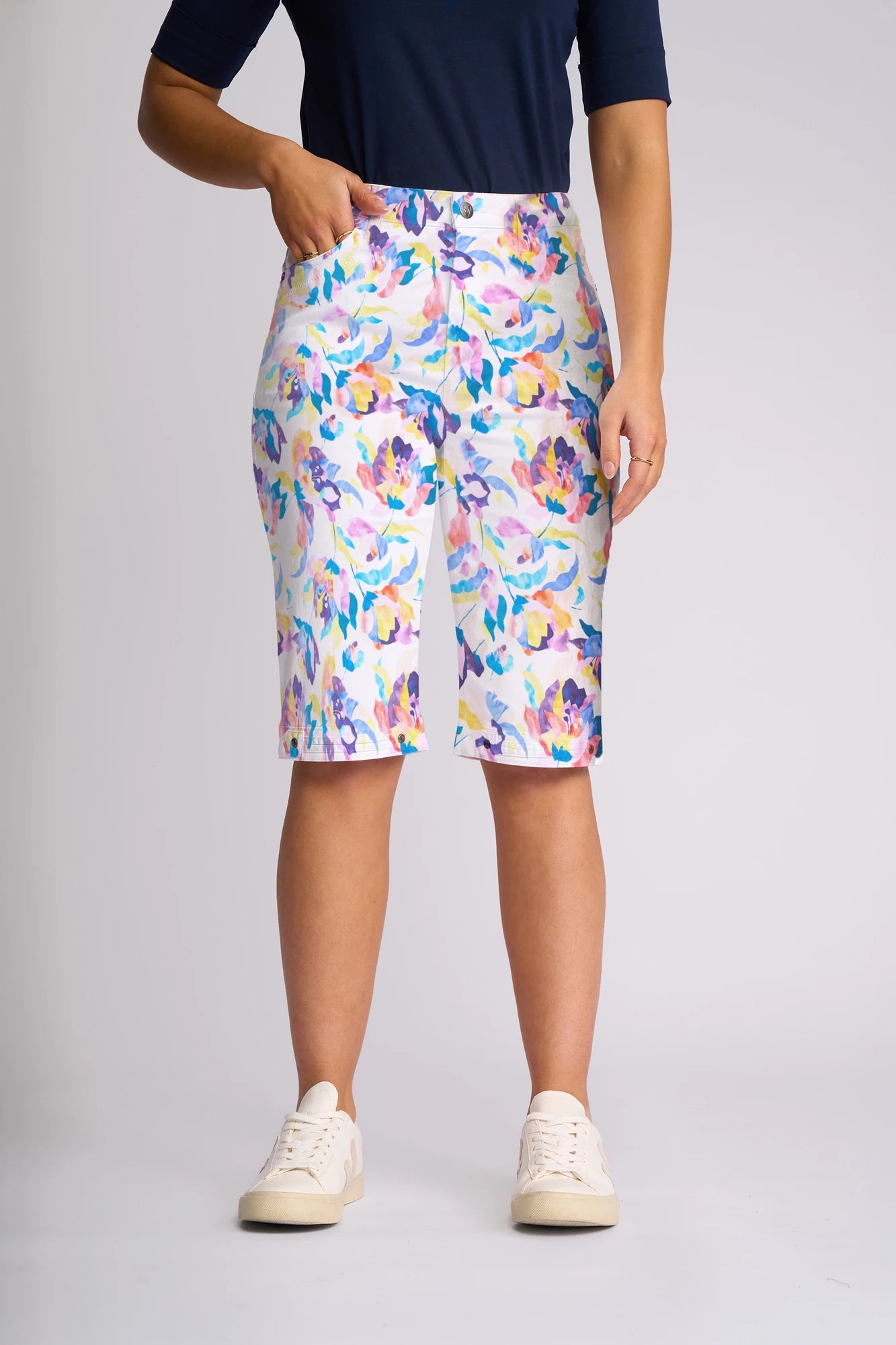 A person wearing the Vassalli Custom Short - Demi, featuring a knee-length, colorful floral print—a true summer staple—pairs it with a navy top and stands with one hand in their pocket. They are also wearing white sneakers.