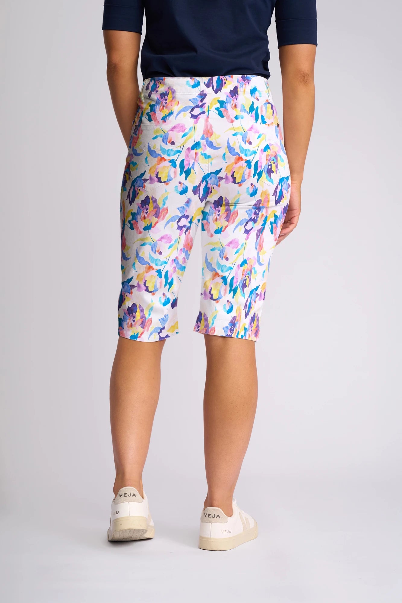 A person wearing Vassalli’s Custom Short - Demi in a vibrant floral print, paired with a navy shirt and white Veja sneakers, stands facing away against a plain light background—showcasing the perfect summer look with these lightweight, breezy shorts as the seasonal staple.