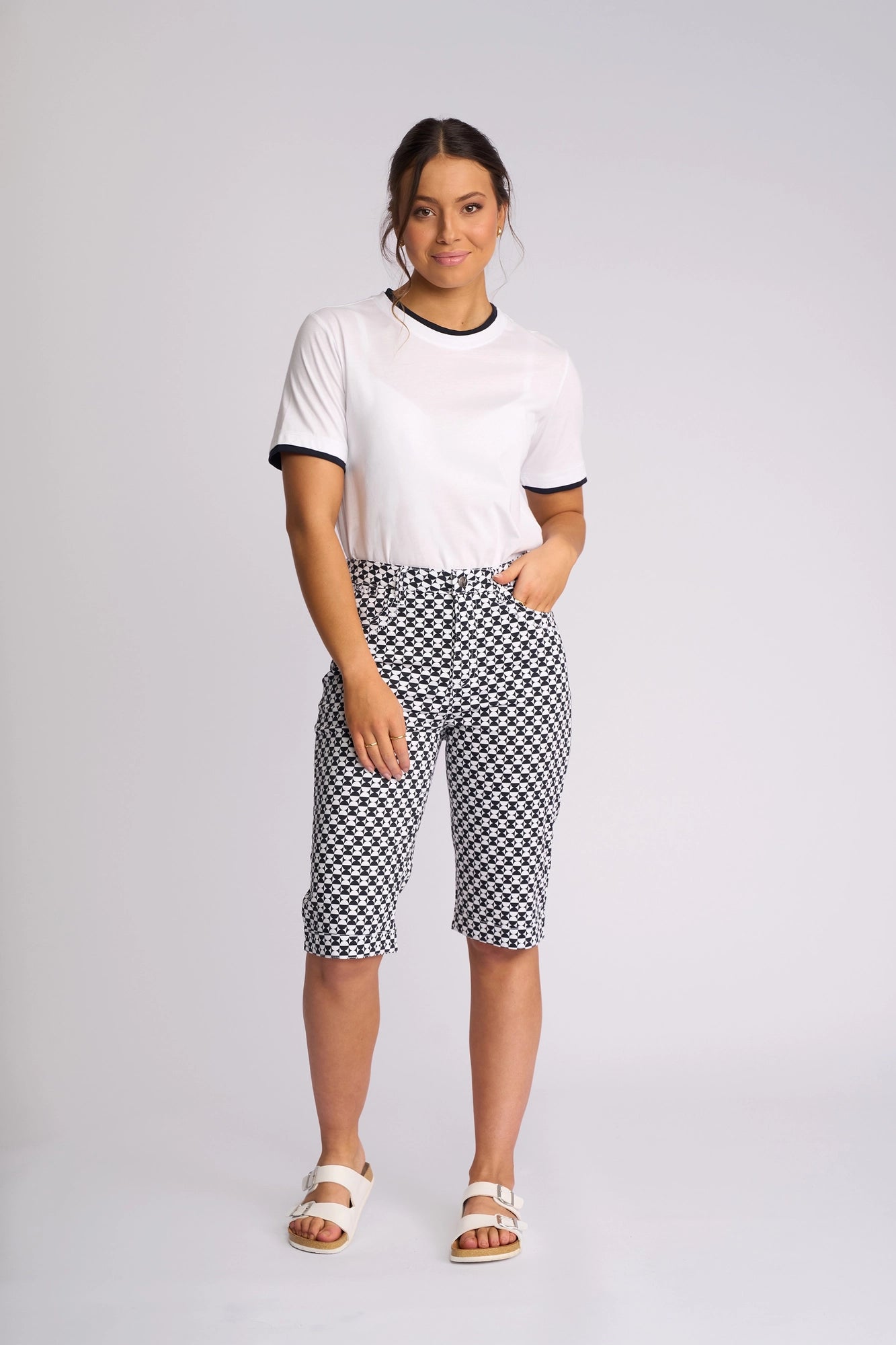 A woman stands against a plain background, wearing the Vassalli Custom Short - Dixie, featuring a black and white geometric print and crafted from a comfortable cotton-elastane blend. She pairs the shorts with a white T-shirt with black trim and white sandals, posing with one hand in her pocket and smiling slightly.