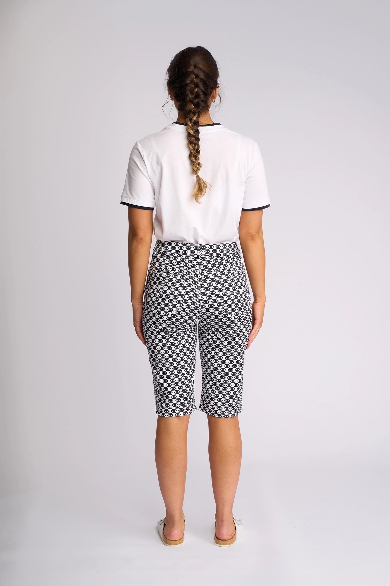 A woman with a long braid stands facing away, wearing a white short-sleeve shirt, the Vassalli Custom Short - Dixie featuring a black-and-white geometric print crafted from a comfortable cotton-elastane blend, and white slip-on shoes, against a plain light background.