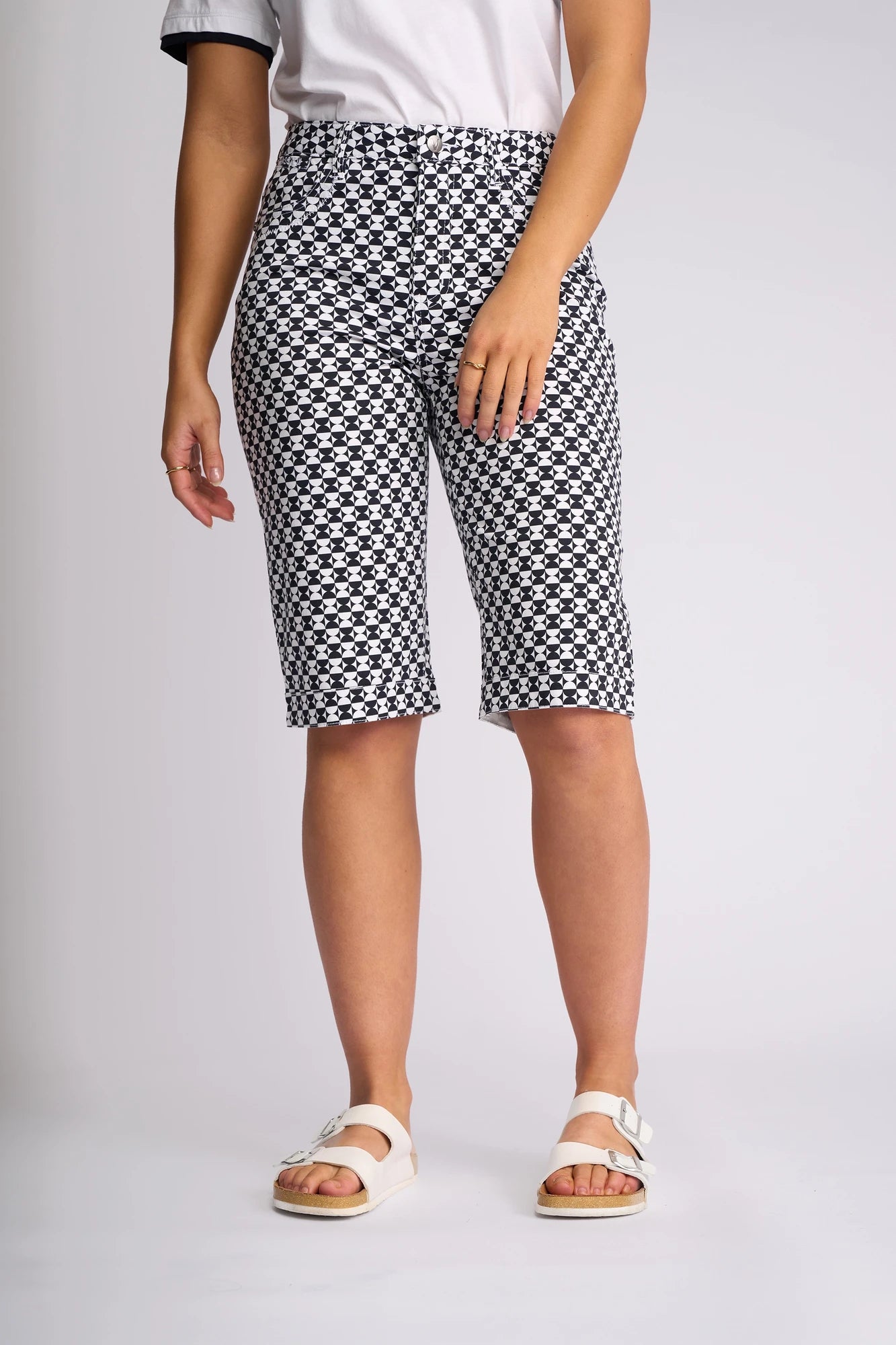 A person dressed in the Vassalli Custom Short - Dixie, featuring a black and white geometric print and knee-length cut in a lightweight cotton-elastane blend, paired with a white t-shirt with dark-edged sleeves and white sandals, stands against a plain light background.