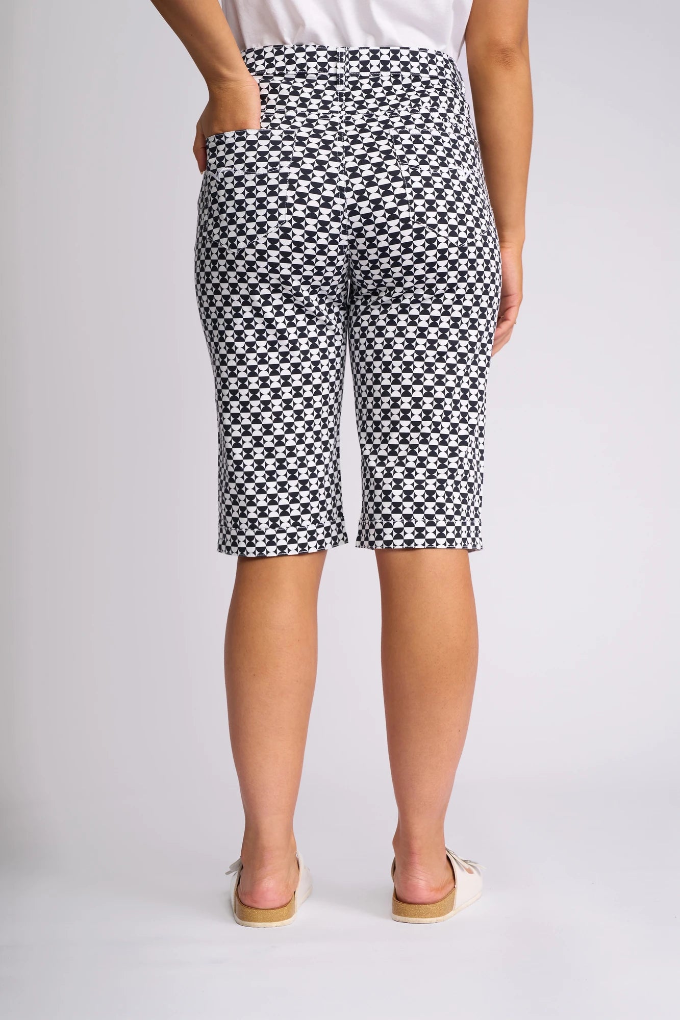 A person stands with their back to the camera, wearing Vassalli’s Custom Short - Dixie in a black and white geometric print, crafted from a comfortable cotton-elastane blend. They pair the knee-length shorts with a white top and light-colored slip-on shoes, one hand tucked into the pocket, posed against a plain background.