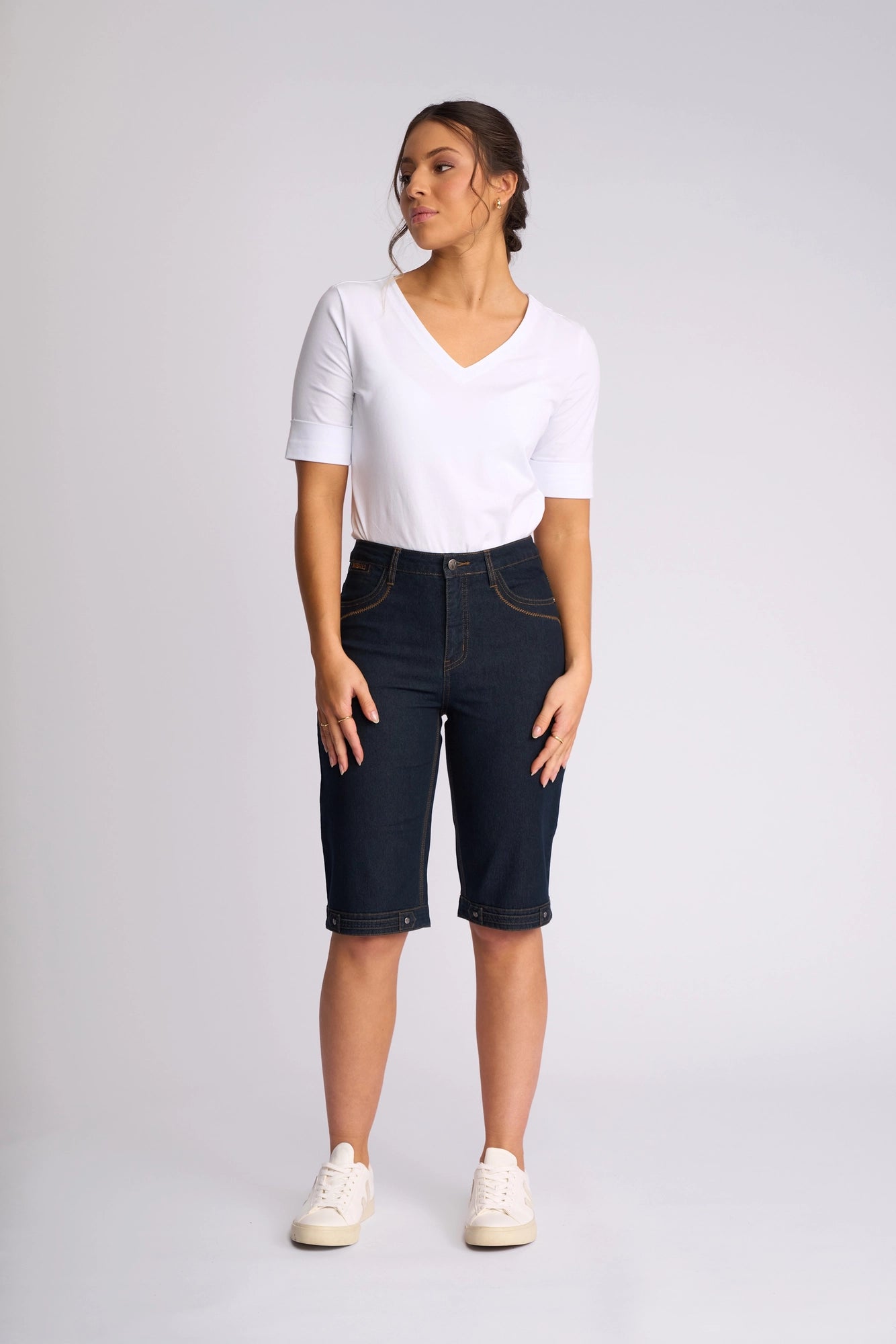 A woman stands against a plain background, wearing a white V-neck top, Vassalli Custom Short in Indigo Denim—crafted from stretch denim for all-day comfort—and white sneakers. She gazes slightly to her left with her hands relaxed by her sides, creating an effortless look perfect for summer dressing.