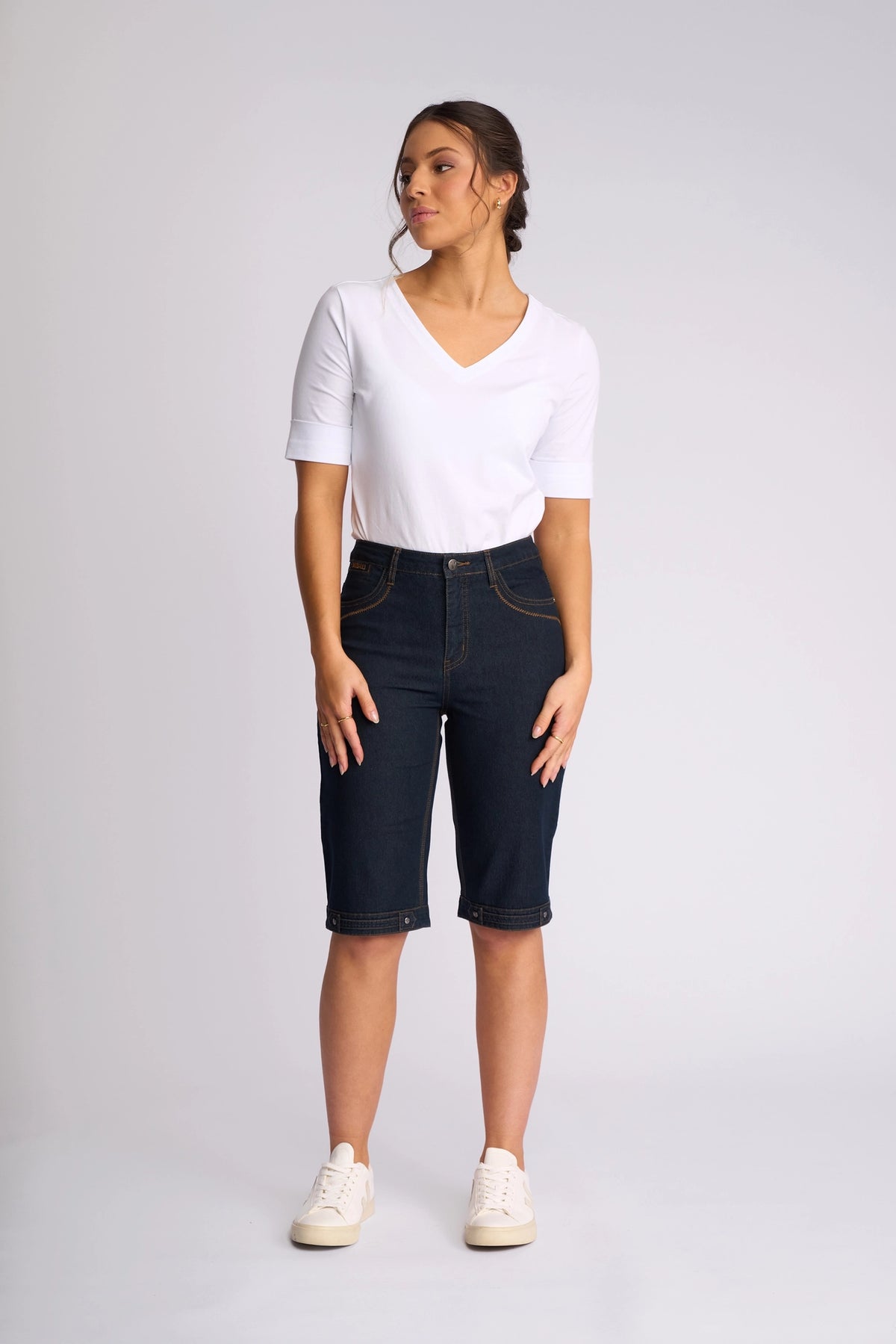 A woman stands against a plain background, wearing a white V-neck top, Vassalli Custom Short in Indigo Denim—crafted from stretch denim for all-day comfort—and white sneakers. She gazes slightly to her left with her hands relaxed by her sides, creating an effortless look perfect for summer dressing.