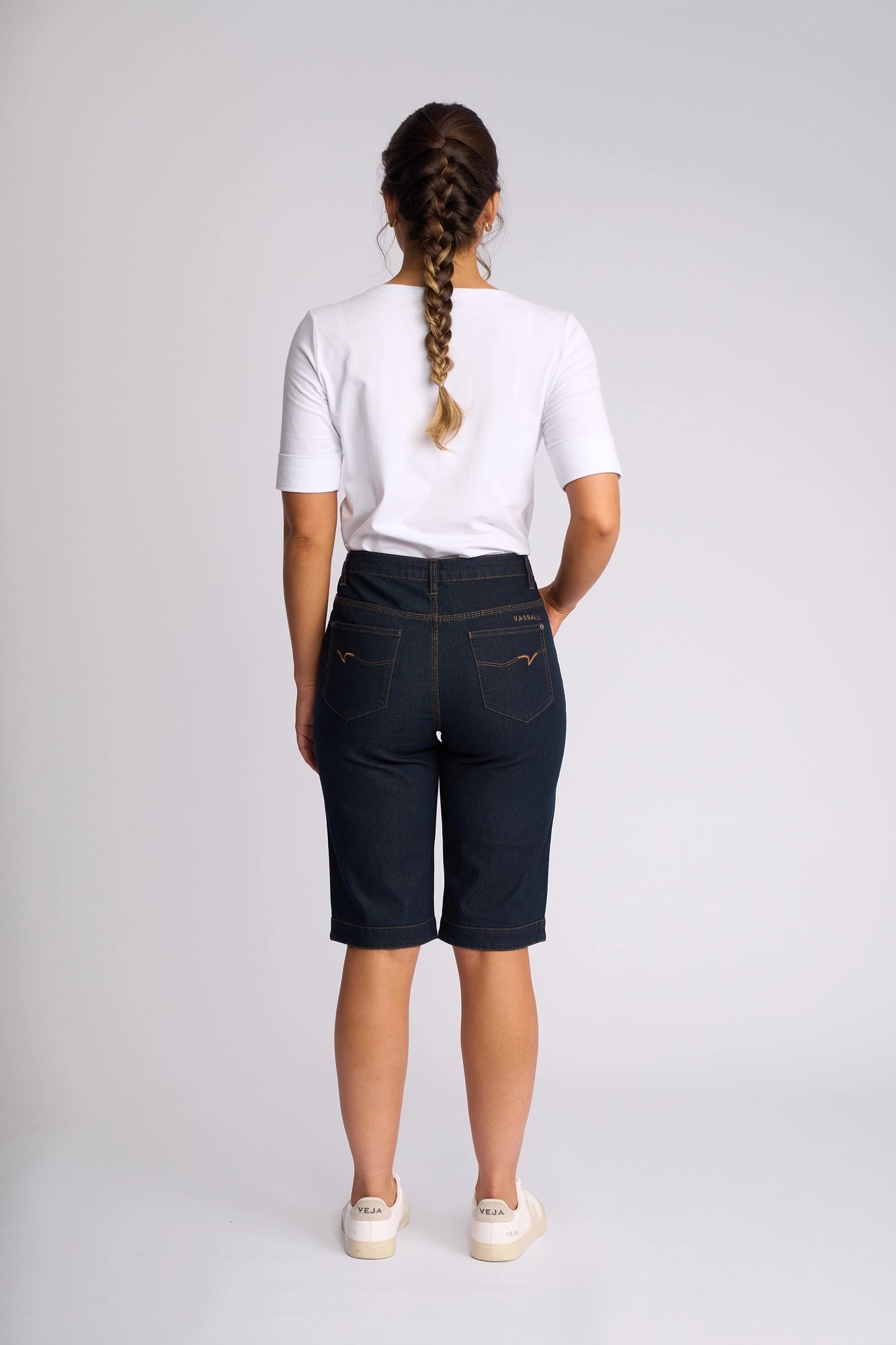 A person with a long braid wears a white t-shirt, Vassalli’s Custom Short in indigo denim crafted from stretch fabric, and white sneakers, standing against a plain light background and facing away from the camera—an effortless look perfect for summer dressing.