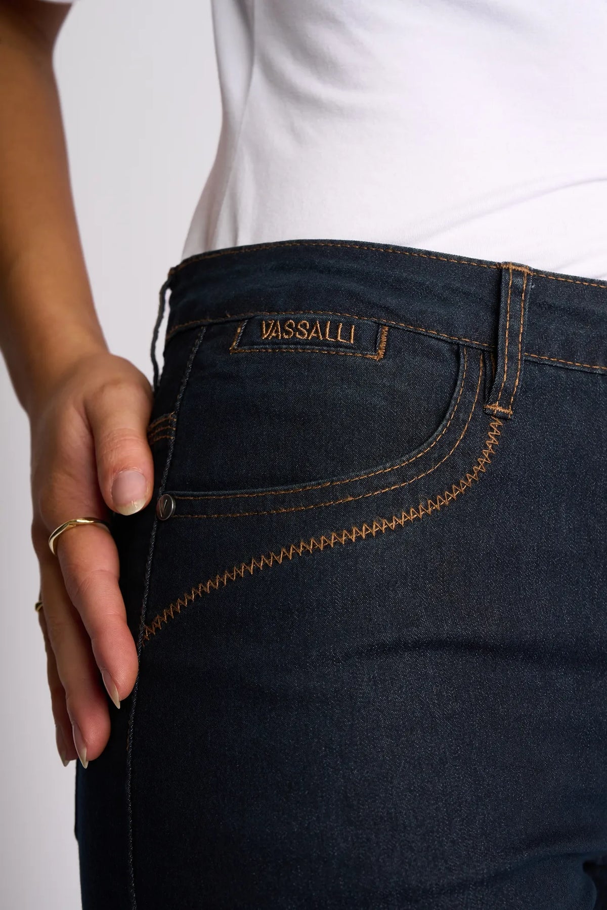 A person wearing Vassalli’s Custom Short in Indigo Denim paired with a white top, with one hand resting near the pocket. Perfect for summer, these shorts feature visible orange stitching and a small label above the pocket.