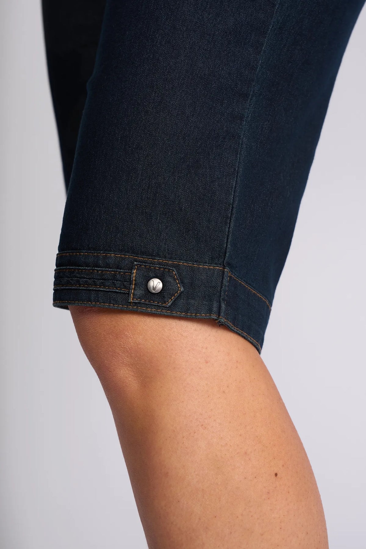 Close-up of a person’s knee wearing the Vassalli Custom Short – Indigo Denim, featuring a decorative buttoned strap detail near the hem—crafted from stretch denim for comfort, perfect for effortless summer dressing.