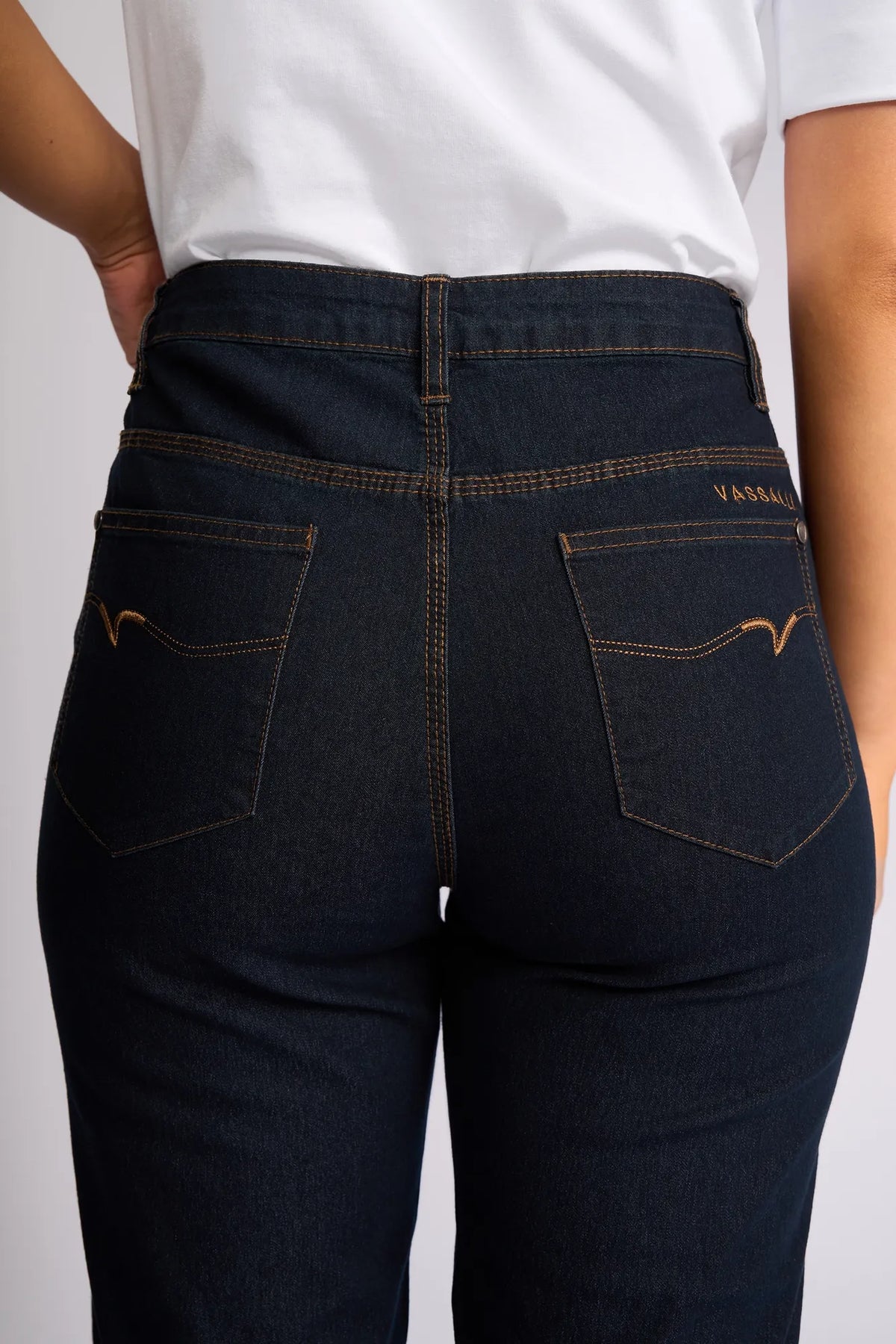 A person, shown from the back, is wearing a white shirt paired with Vassalli’s Custom Short in Indigo Denim. Crafted from stretch denim for added comfort, these shorts feature two back pockets accented with brown stitching and a small brand label above the right pocket—perfect for effortless summer dressing.