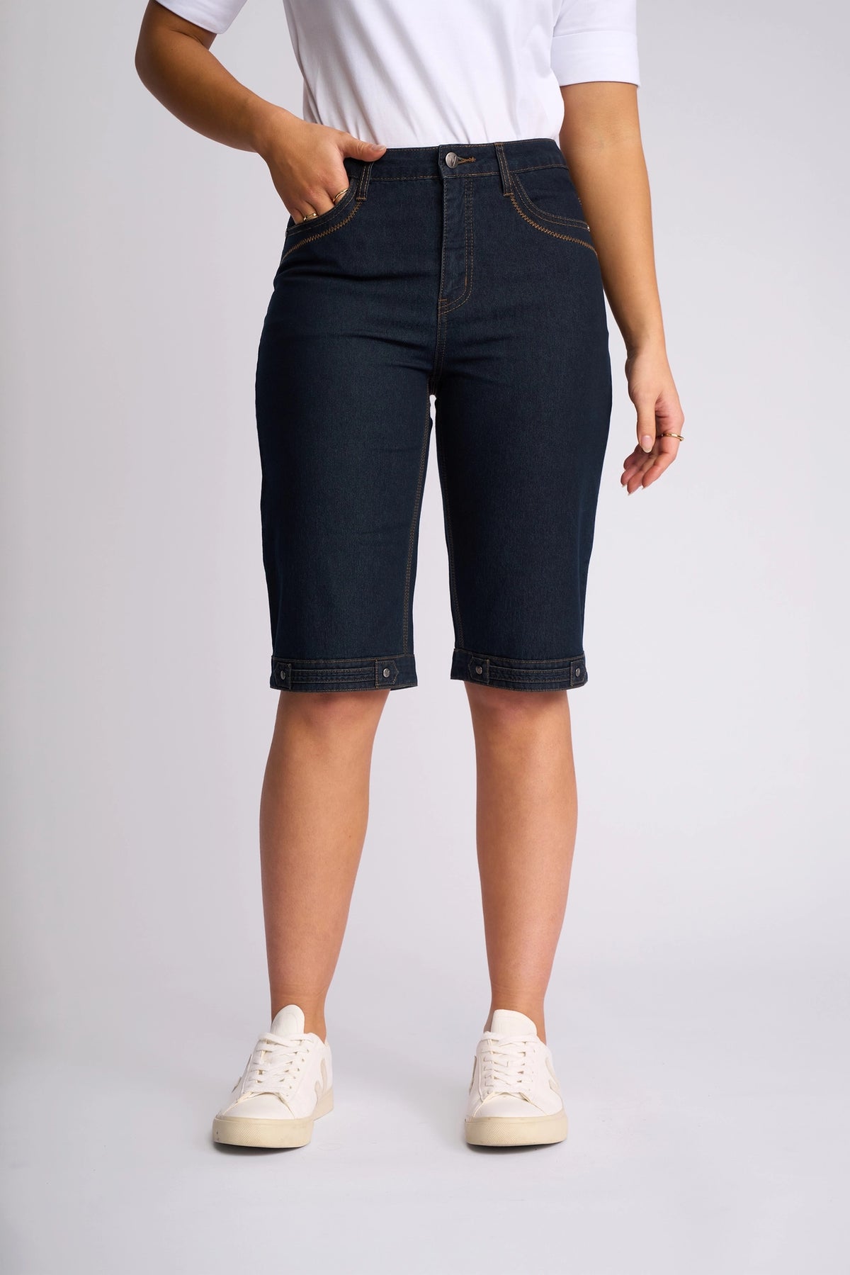 A person is wearing a white shirt, Vassalli Custom Short in indigo denim crafted from comfortable stretch fabric, and white sneakers. Perfect for summer dressing, they are standing with one hand in a pocket against a plain light background.